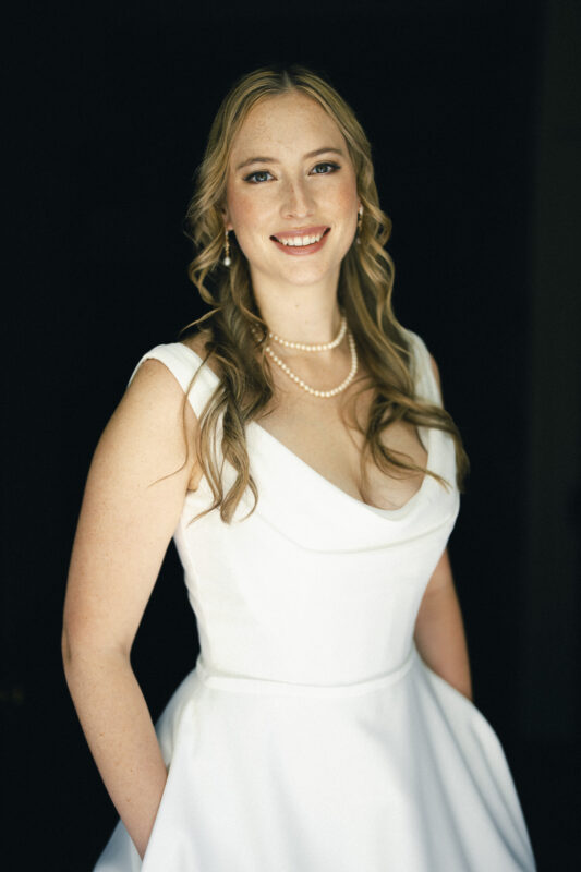 A bride in a white dress with curled hair and pearl necklaces smiles against a dark background.