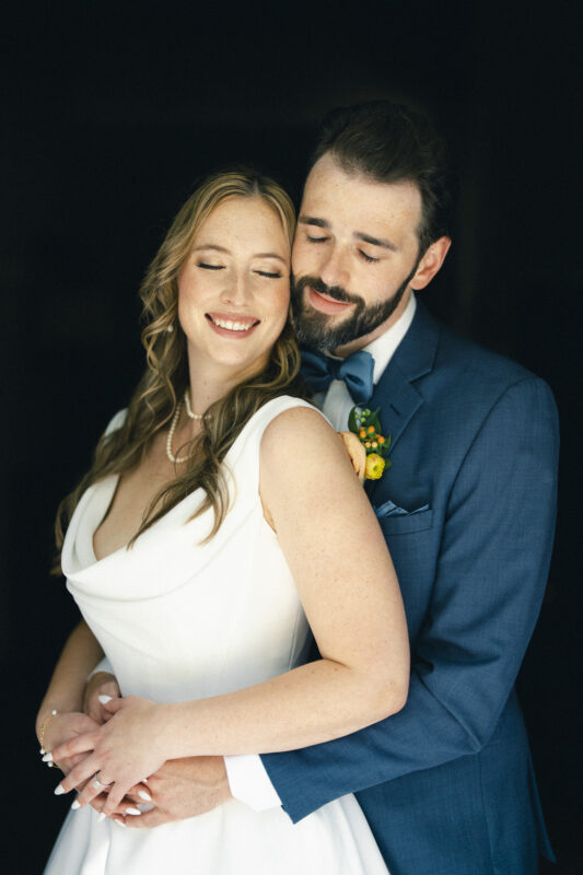 A bride and groom embrace lovingly. The bride wears a white dress with a pearl necklace, and the groom is in a blue suit with a bow tie and boutonniere. Both have eyes closed, smiling softly against a dark background.