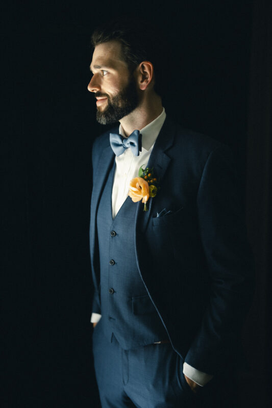 A man in a dark blue suit and bow tie stands in soft lighting against a dark background. He has a beard and is looking to the left, with hands in his pockets. A light-colored flower boutonniere is pinned to his suit.