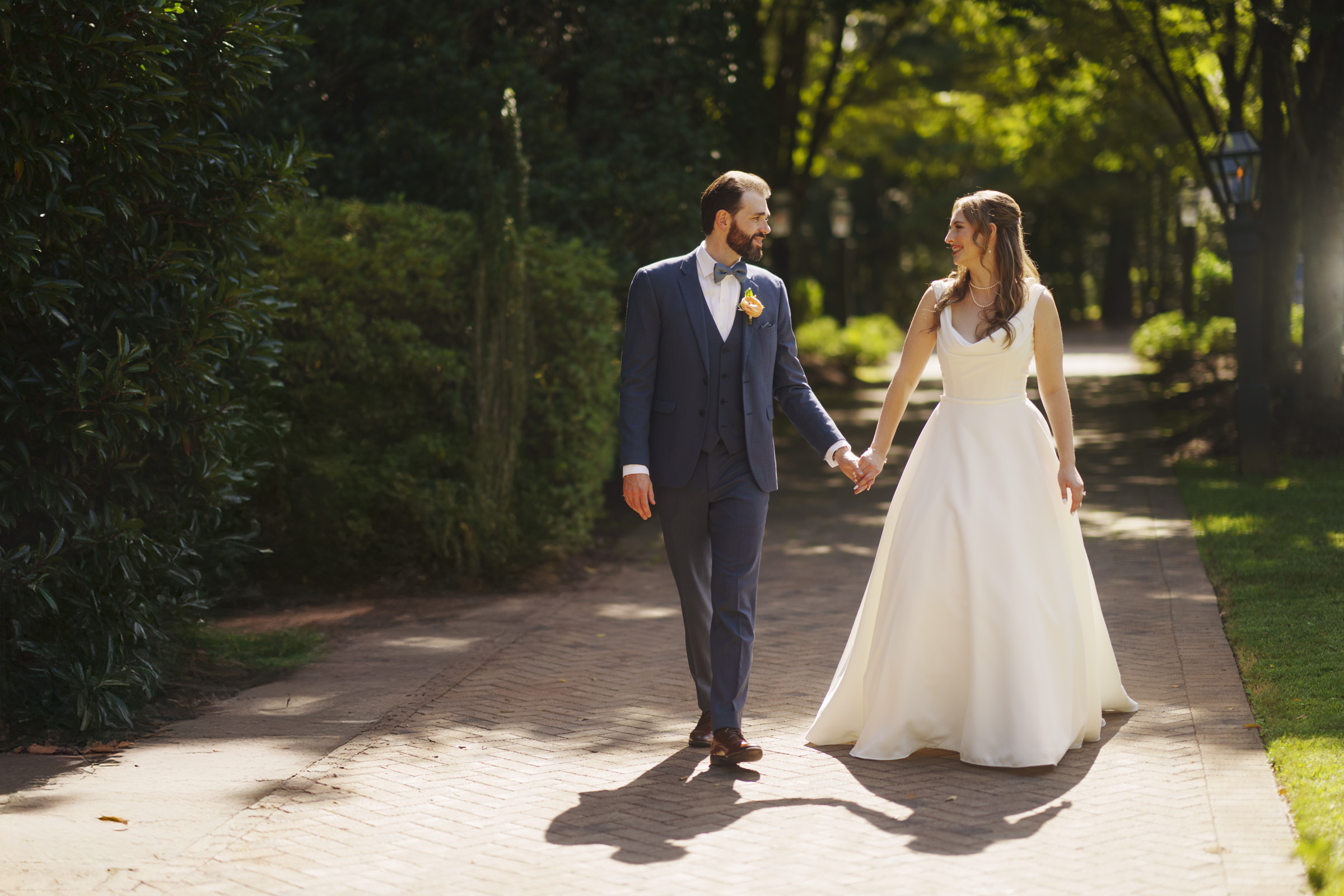 A bride and groom walk hand in hand down a sunlit path surrounded by greenery. The groom is in a blue suit with a boutonniere, and the bride wears a flowing white gown, both smiling at each other.