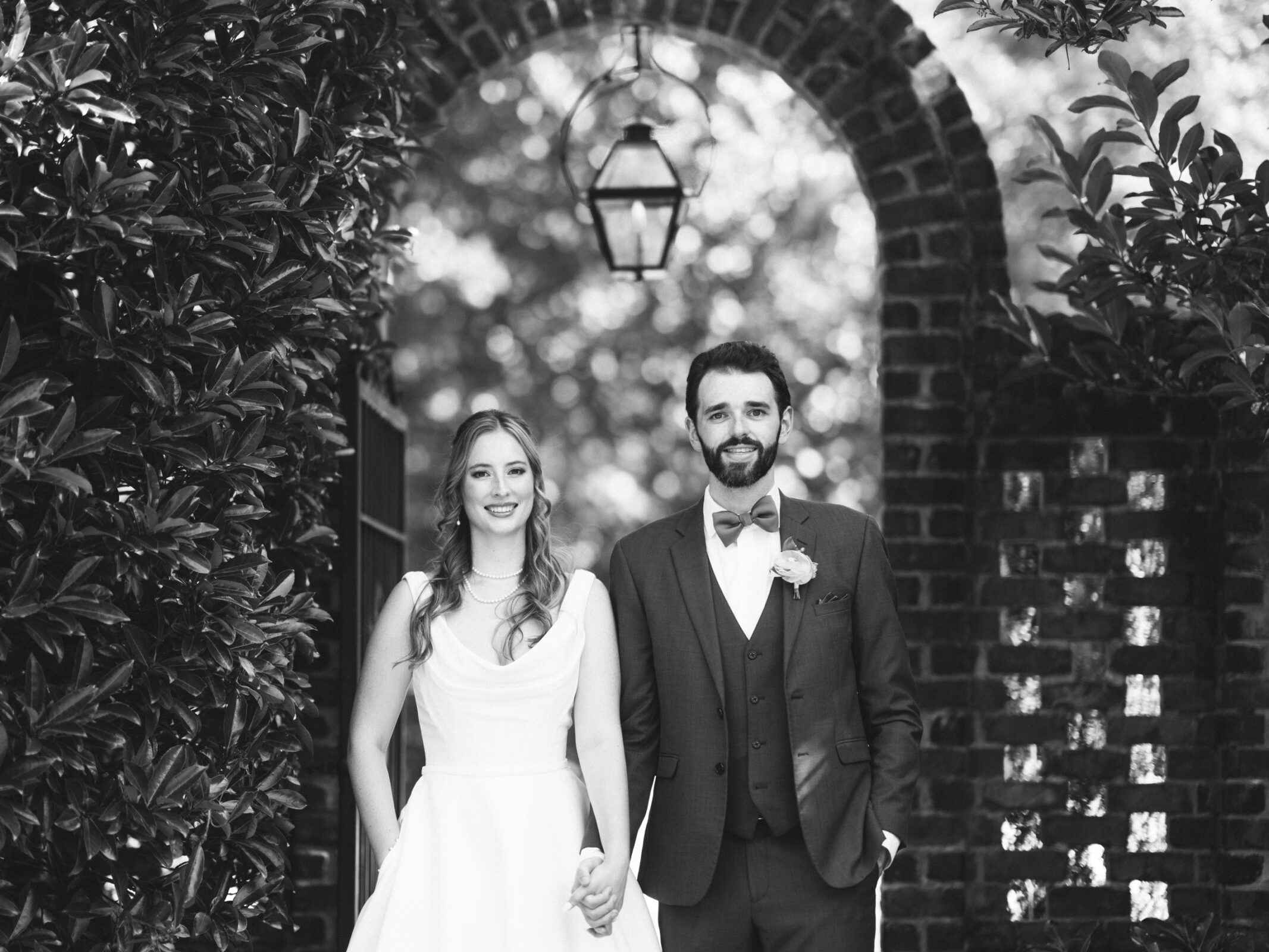 A bride and groom stand hand in hand under a brick archway in a lush garden. The bride wears a simple white dress, and the groom is in a suit with a bow tie. Both are smiling. The photo is in black and white.