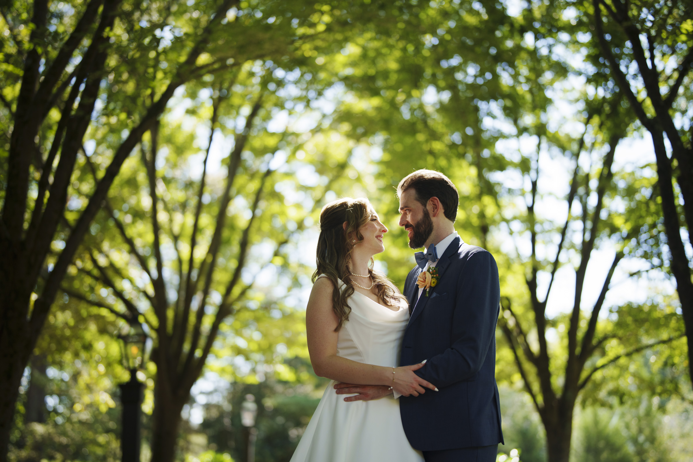 A bride and groom stand facing each other, smiling, under a canopy of sunlit trees. The bride wears a white dress, and the groom is in a blue suit with a boutonniere. The background is lush and green, adding a serene vibe to the scene.