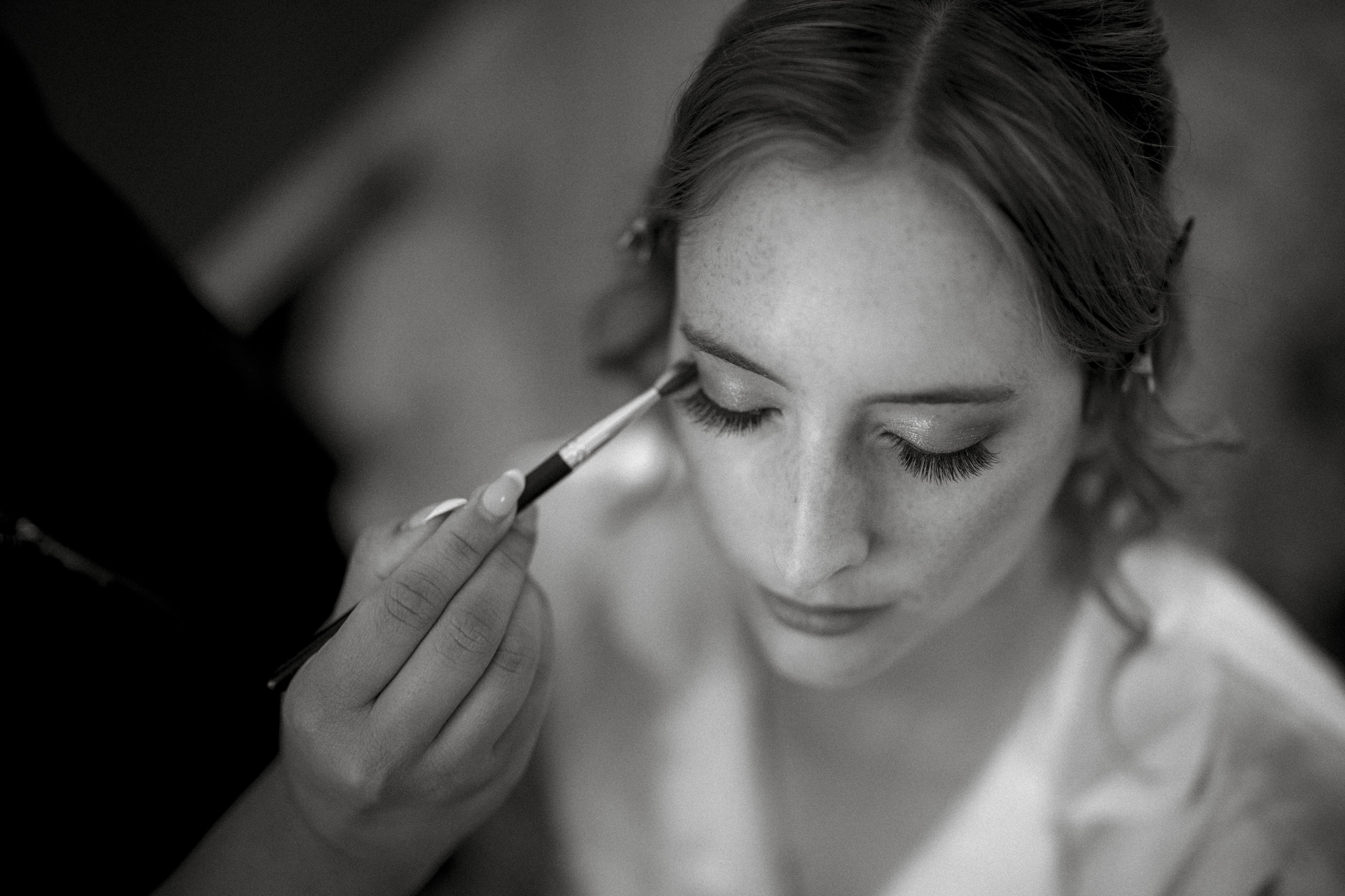 A black and white image of a person with closed eyes having makeup applied with a brush. Hair is pinned back, and the focus is on their serene expression and the makeup artists hand.