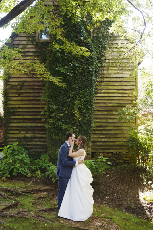 A couple in wedding attire shares a kiss in front of a rustic wooden building covered with ivy. They are surrounded by lush greenery and trees. The groom is wearing a blue suit, and the bride is in a white dress.