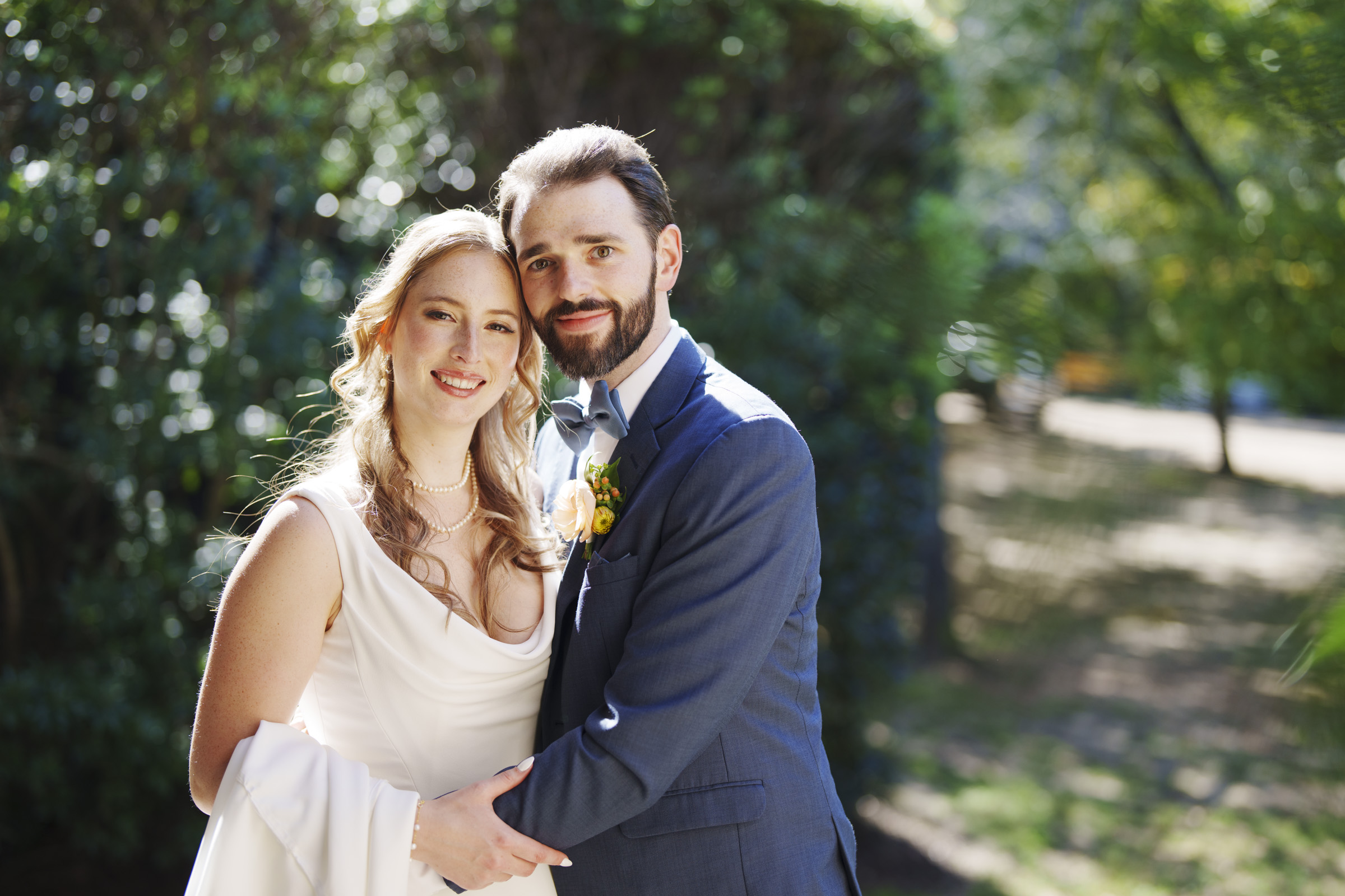 A bride and groom stand outdoors in a sunlit garden. The bride is in a white dress and the groom is in a blue suit with a bow tie and boutonniere. They are smiling and embracing each other. Lush greenery is visible in the background.