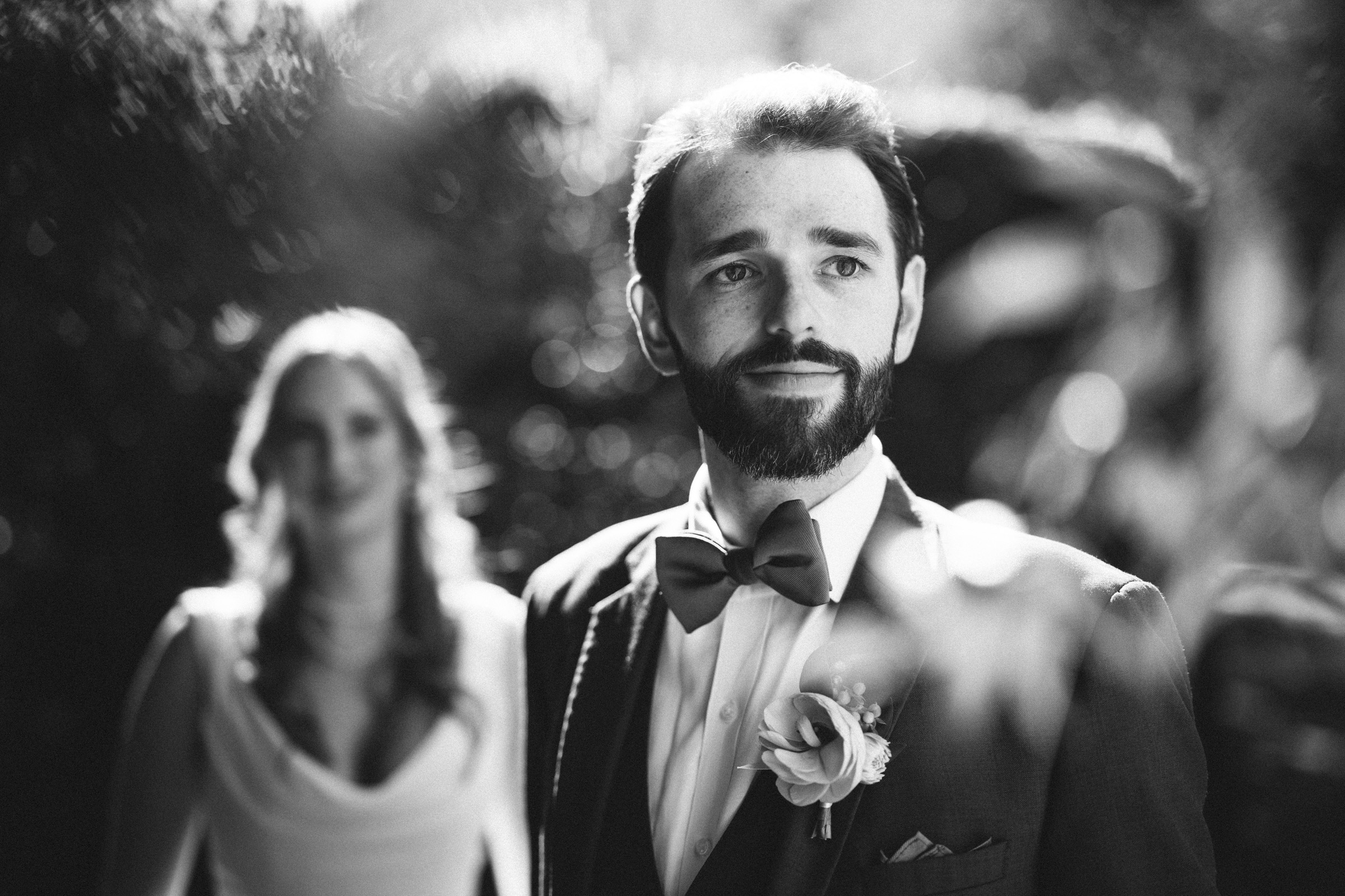 Black and white photo of a man in a suit with a bow tie and boutonniere, looking thoughtfully to the side. A woman in a dress stands behind him, slightly out of focus. The background is blurred, suggesting an outdoor setting.