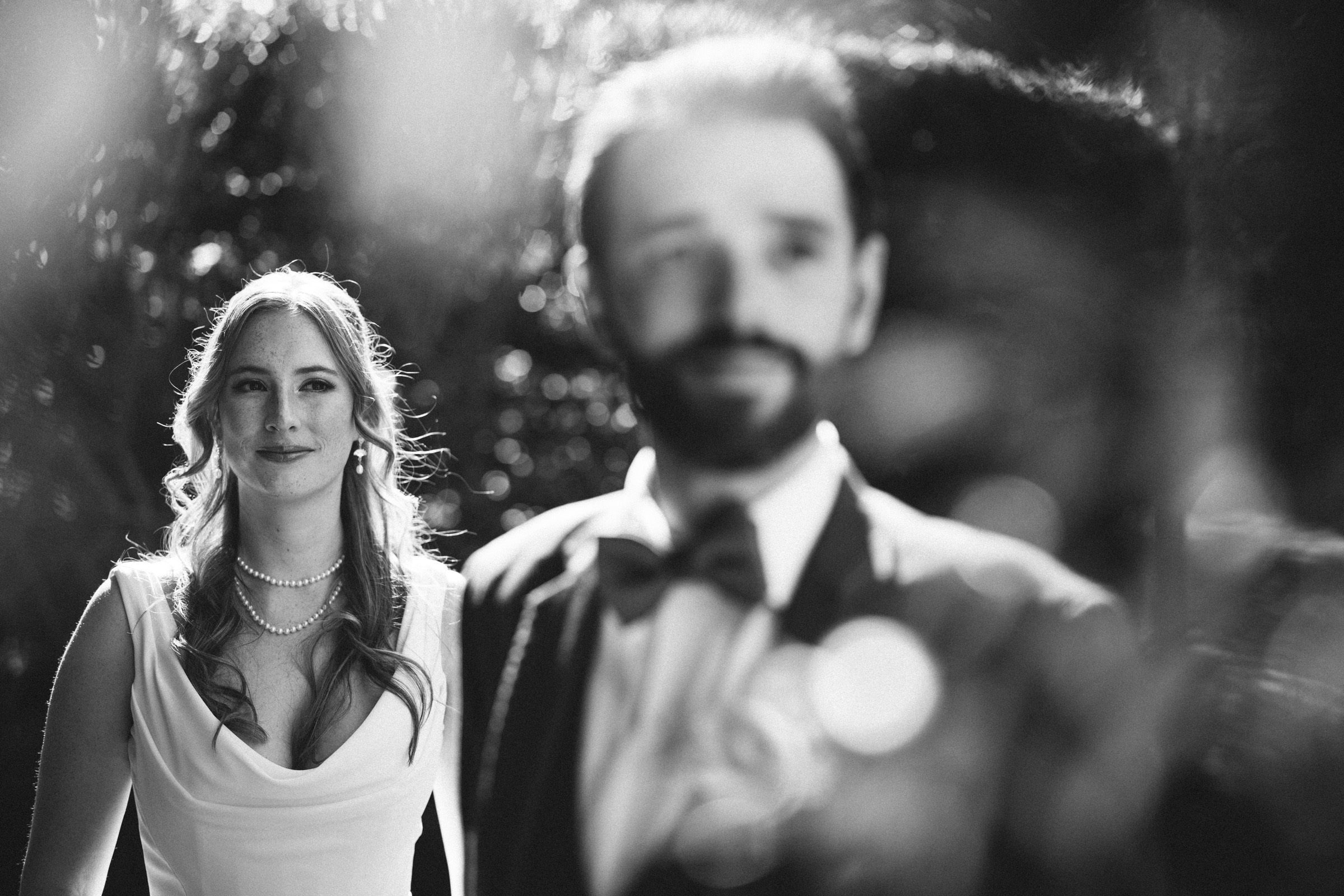 A black and white photo of a wedding couple. The woman is wearing a dress with a pearl necklace, smiling softly in the background. The man, in focus, wears a suit and bow tie, looking to the side. Sunlight filters through the trees.