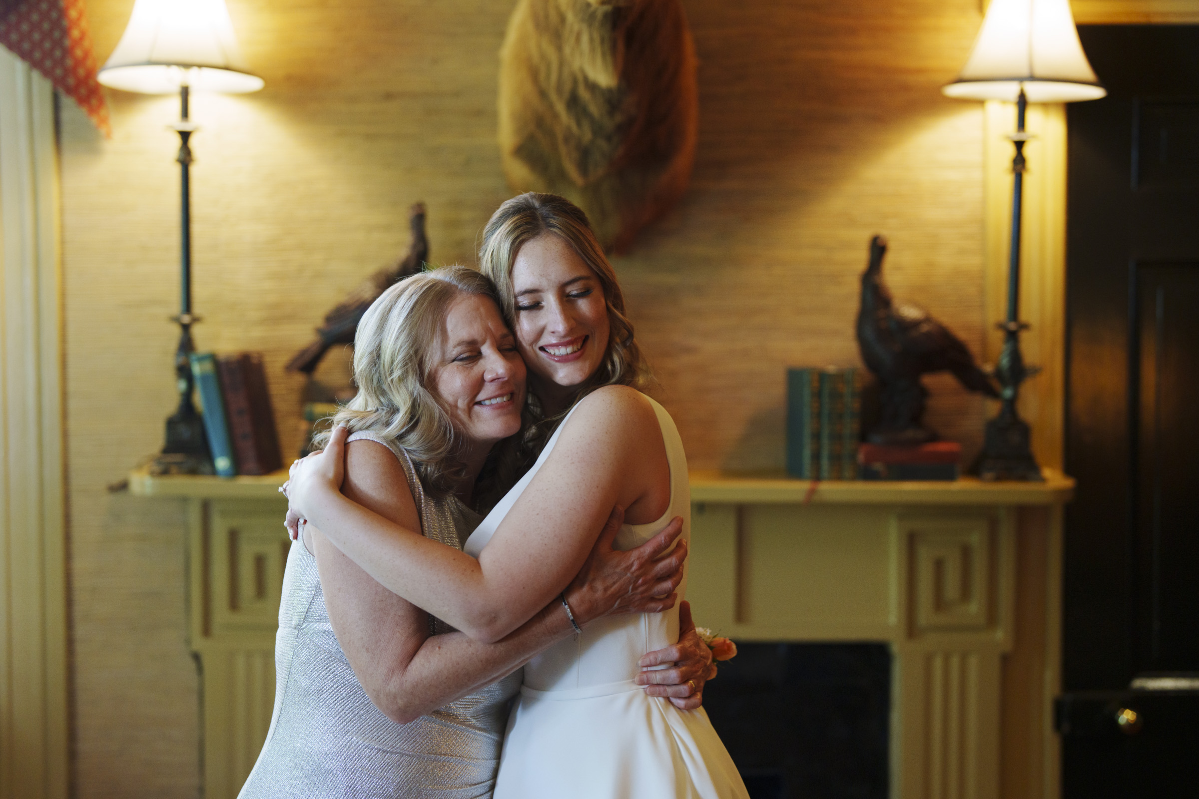 A woman in a white dress hugs an older woman in a light-colored dress, both smiling warmly. They stand in a cozy room with two lamps, decorative books, and figurines on a mantel. The mood is joyful and intimate.