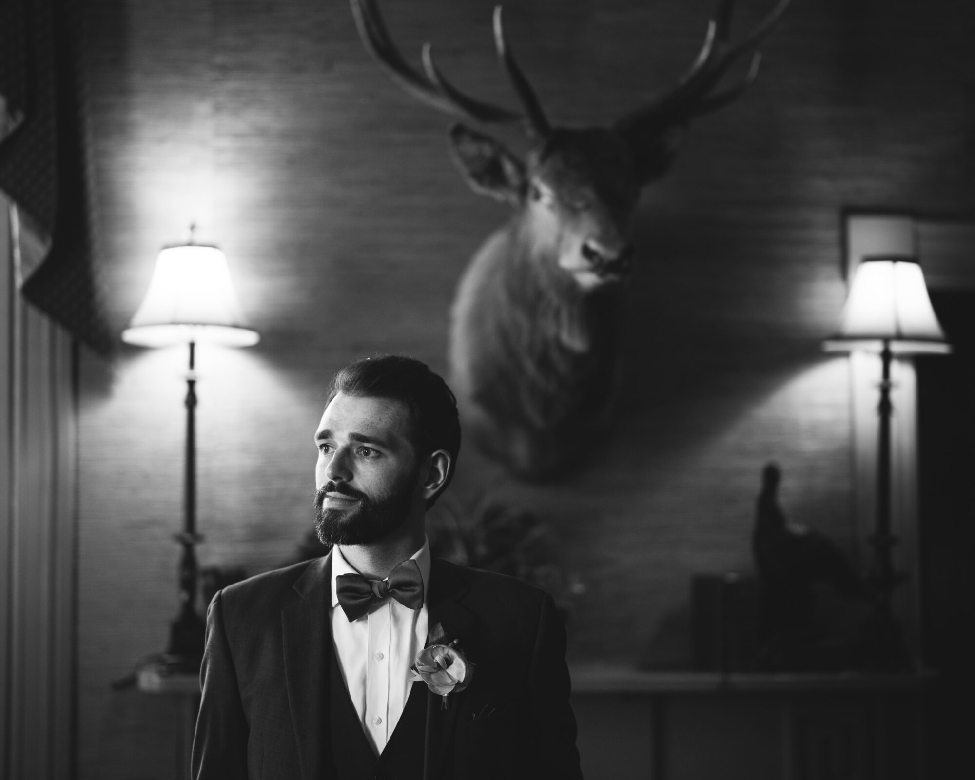 A groom with a beard stands in a room, wearing a suit with a bow tie and boutonniere. Two lamps flank him, and a mounted deer head is visible on the wall behind. The image is in black and white.