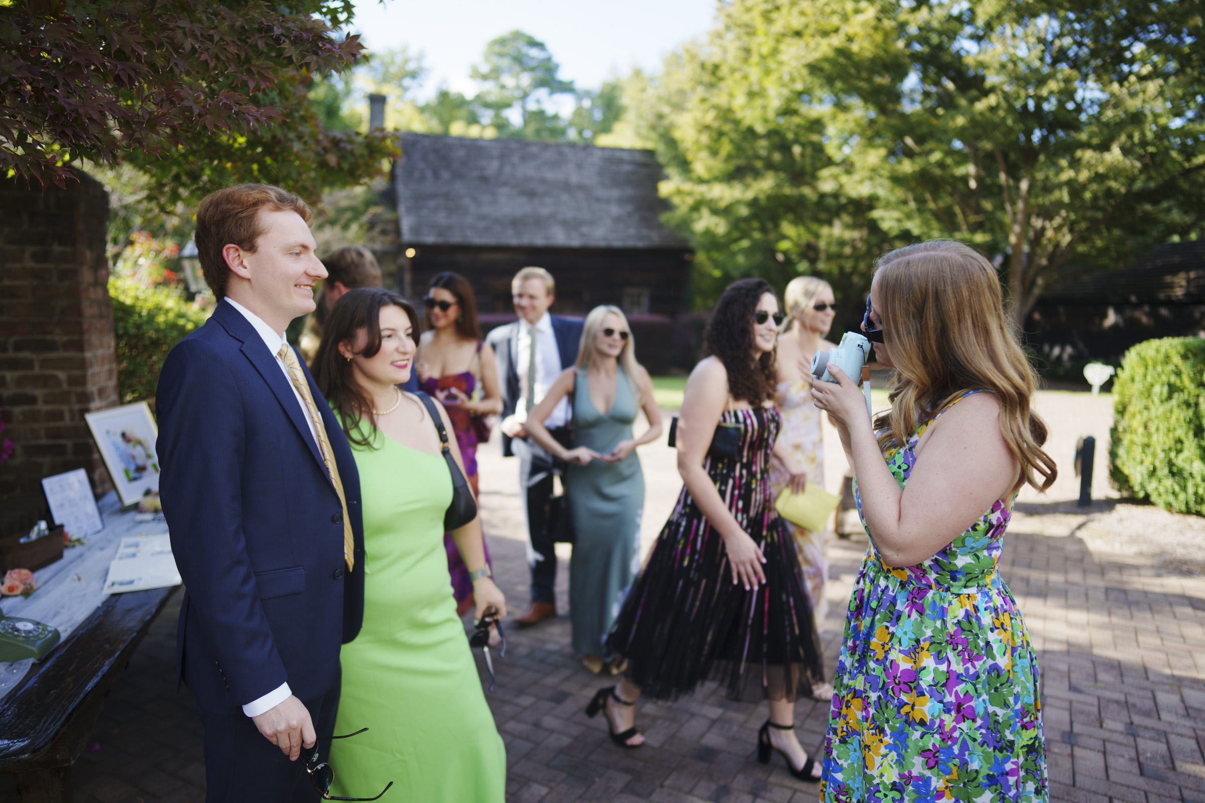 A group of people in formal attire are gathered outdoors. A woman in a floral dress is taking a photo of a man in a suit and a woman in a green dress. Others are standing and smiling in the background among trees and brick paths.