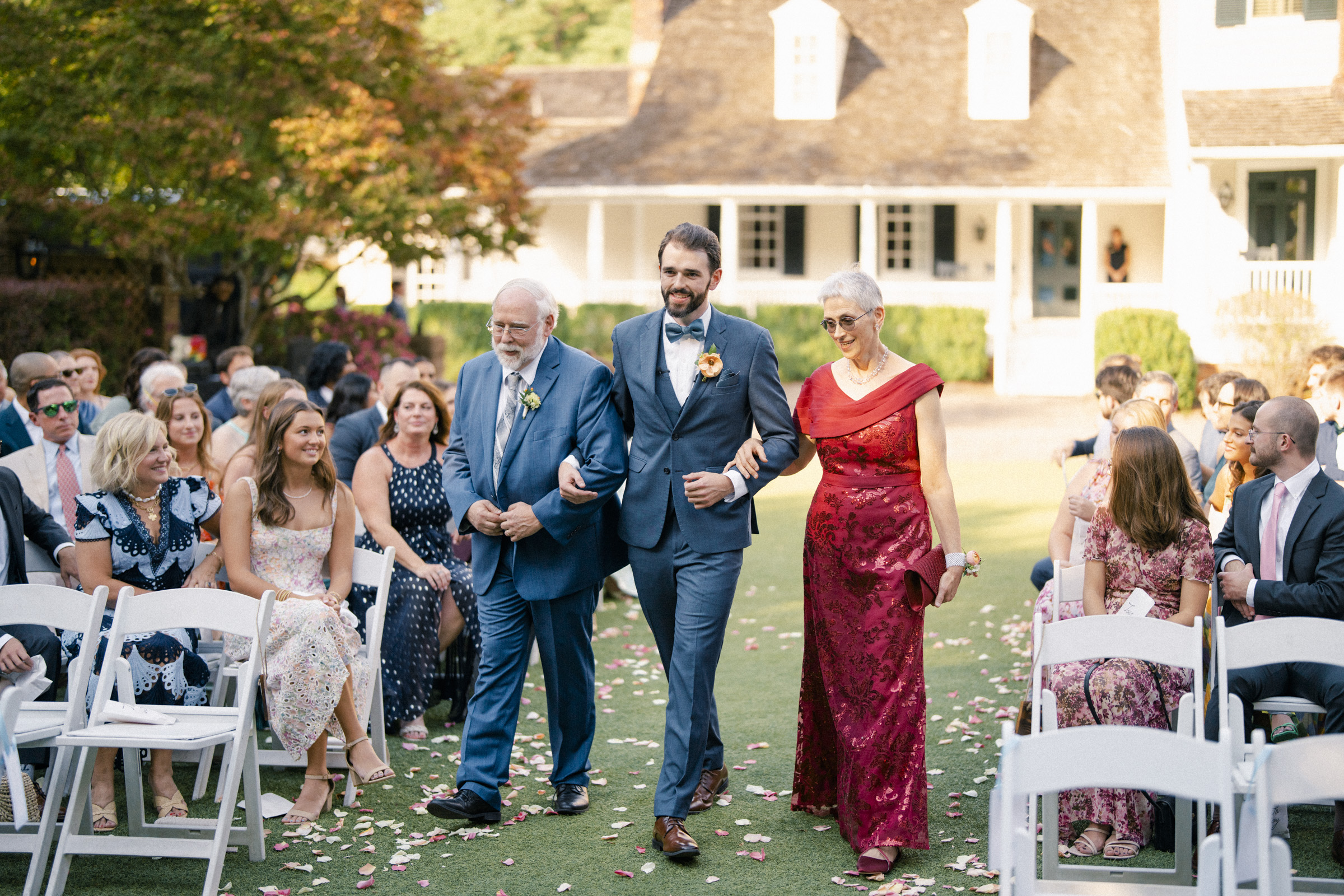 A bearded man in a blue suit walks down an outdoor wedding aisle, smiling, with an elderly man in a blue suit on his left and a woman in a red dress on his right. Guests seated on white chairs watch them. Scattered petals line the aisle.