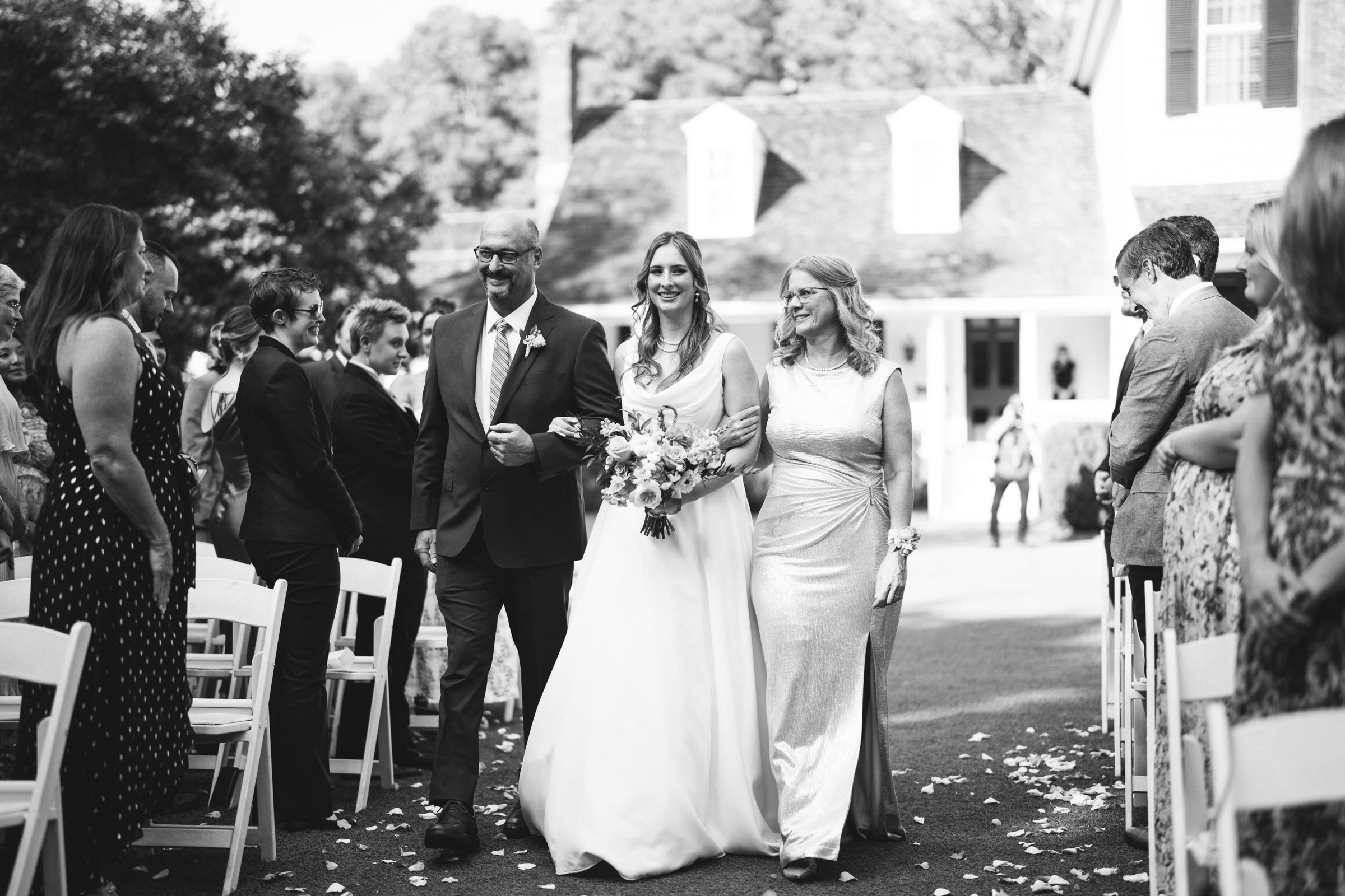 A bride in a white gown walks down the aisle, holding a bouquet, escorted by a man and woman. Guests seated on white chairs look on, smiling. Petals are scattered on the path. The setting is outdoors, beside a house. The photo is in black and white.