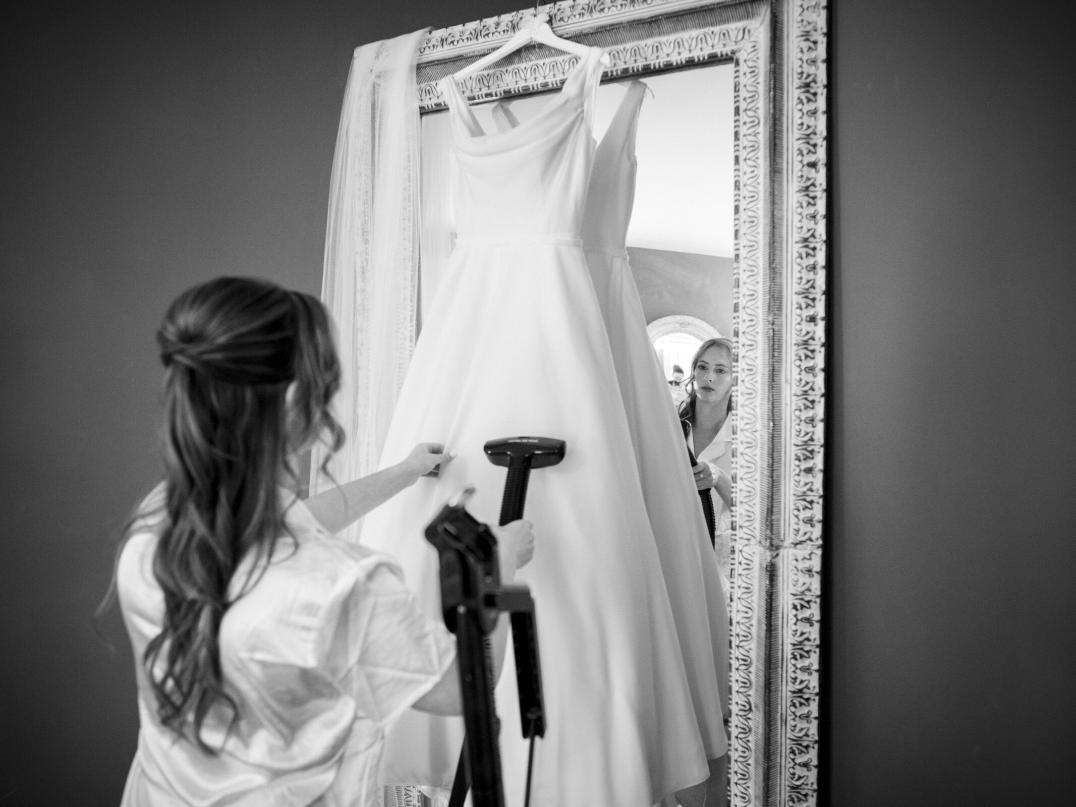 A woman with long hair uses a steamer on a white wedding dress hanging in front of a large mirror. A veil is draped over the top of the mirror. The scene is black and white, reflecting attention to wedding preparations.