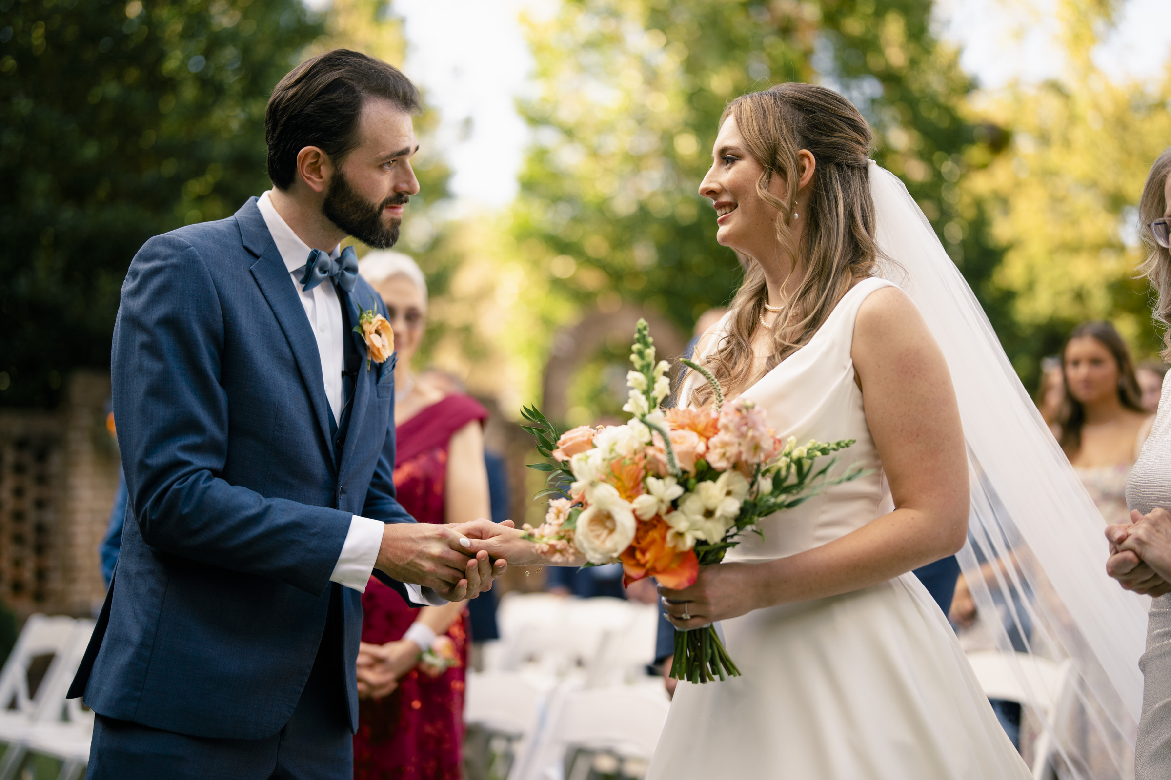 A bride in a white dress and veil holds a bouquet of flowers, smiling at a groom in a blue suit and bow tie. They are outdoors with trees in the background, surrounded by guests seated in white chairs.