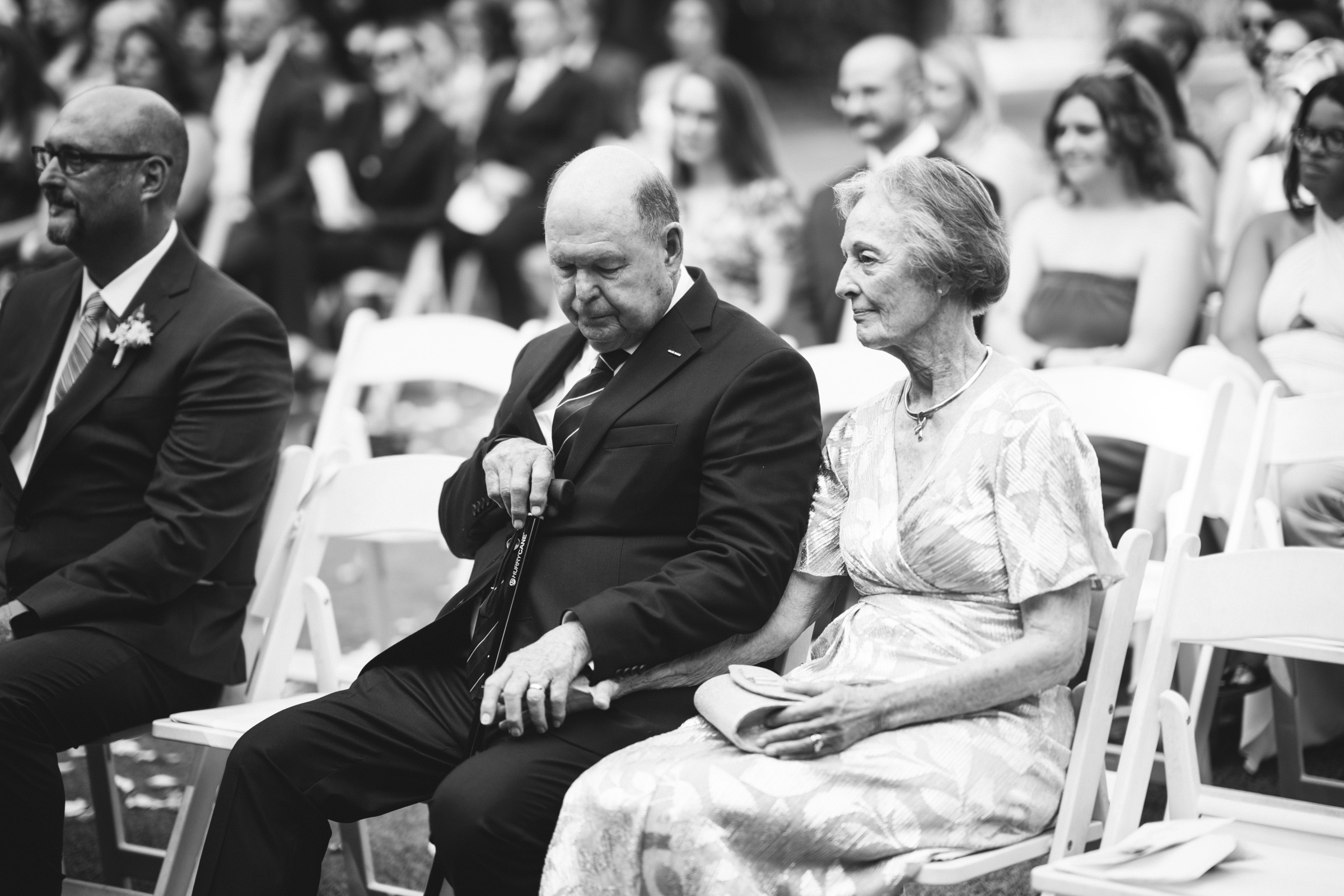 An elderly couple sits on white chairs at an outdoor event. The man wears a suit and holds a cane, while the woman wears a patterned dress. They are surrounded by other attendees in formal attire. The mood is calm and focused on the event happening.