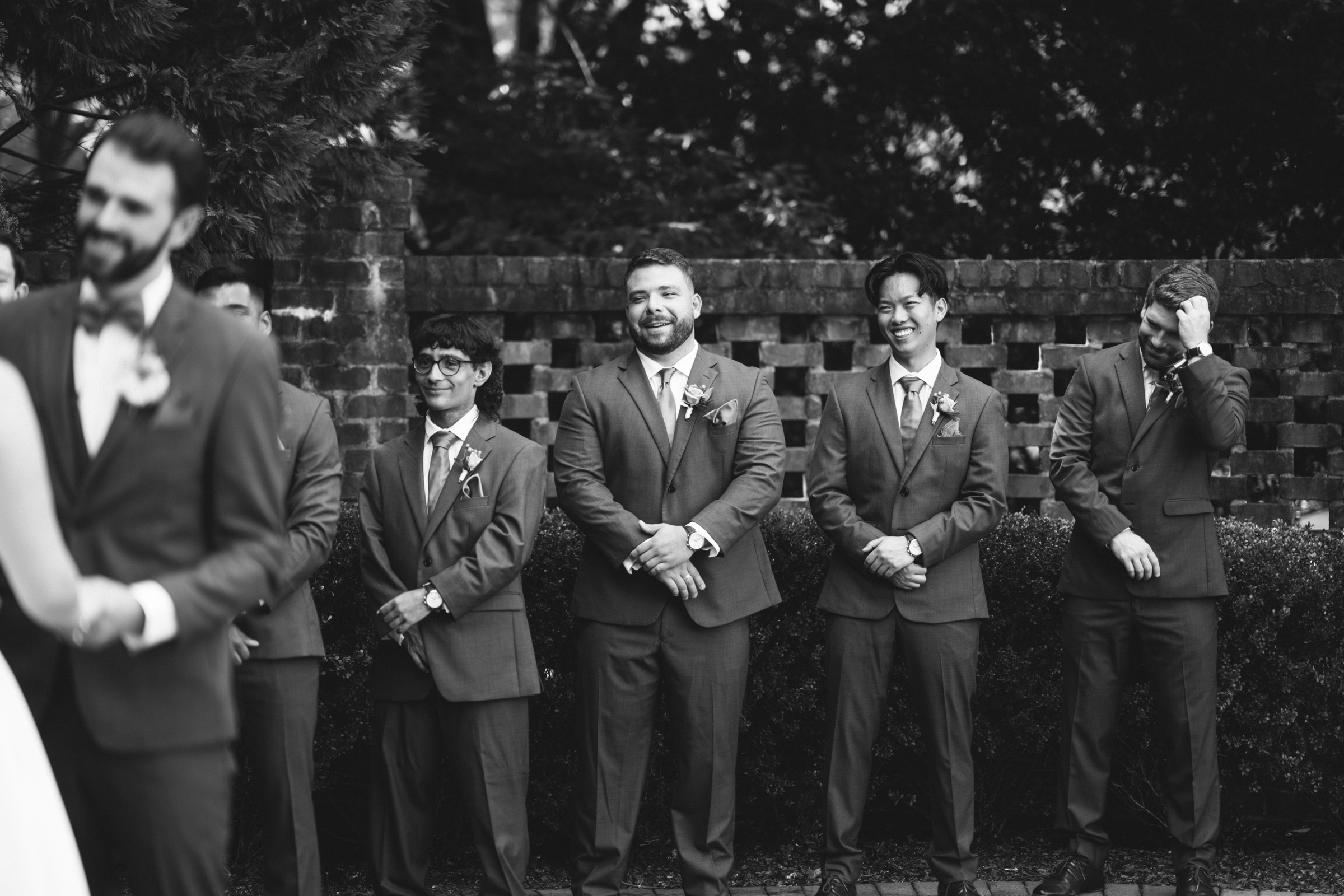 A black and white photo shows a group of groomsmen standing outside. They are dressed in suits with boutonnieres. The background features a brick wall and foliage. Some groomsmen are smiling while looking toward a person in the foreground.