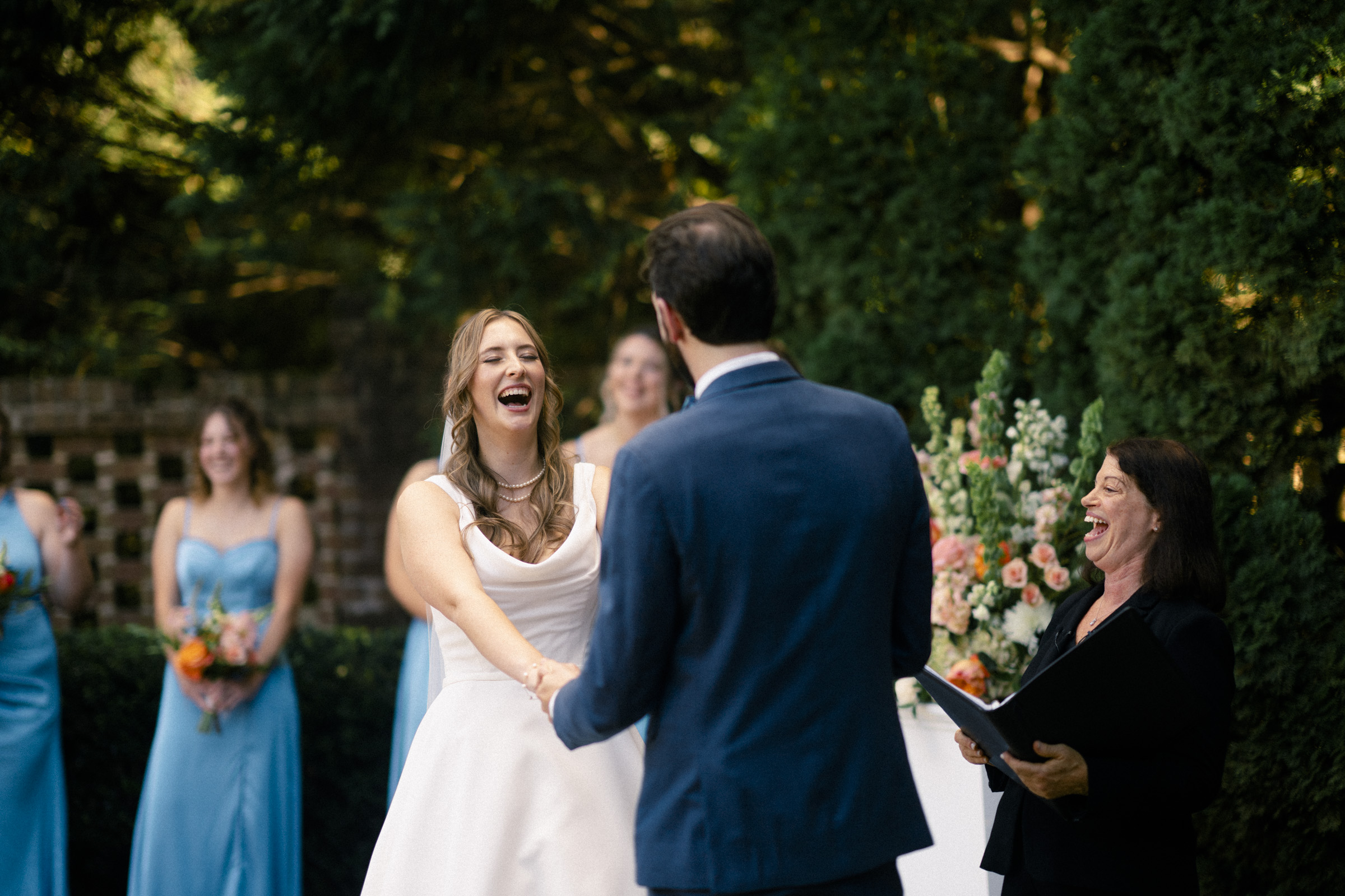 A bride and groom are holding hands and smiling during an outdoor wedding ceremony. The bride is in a white dress, and the groom is in a blue suit. Bridesmaids in blue dresses stand nearby, and an officiant is present. Lush greenery surrounds them.