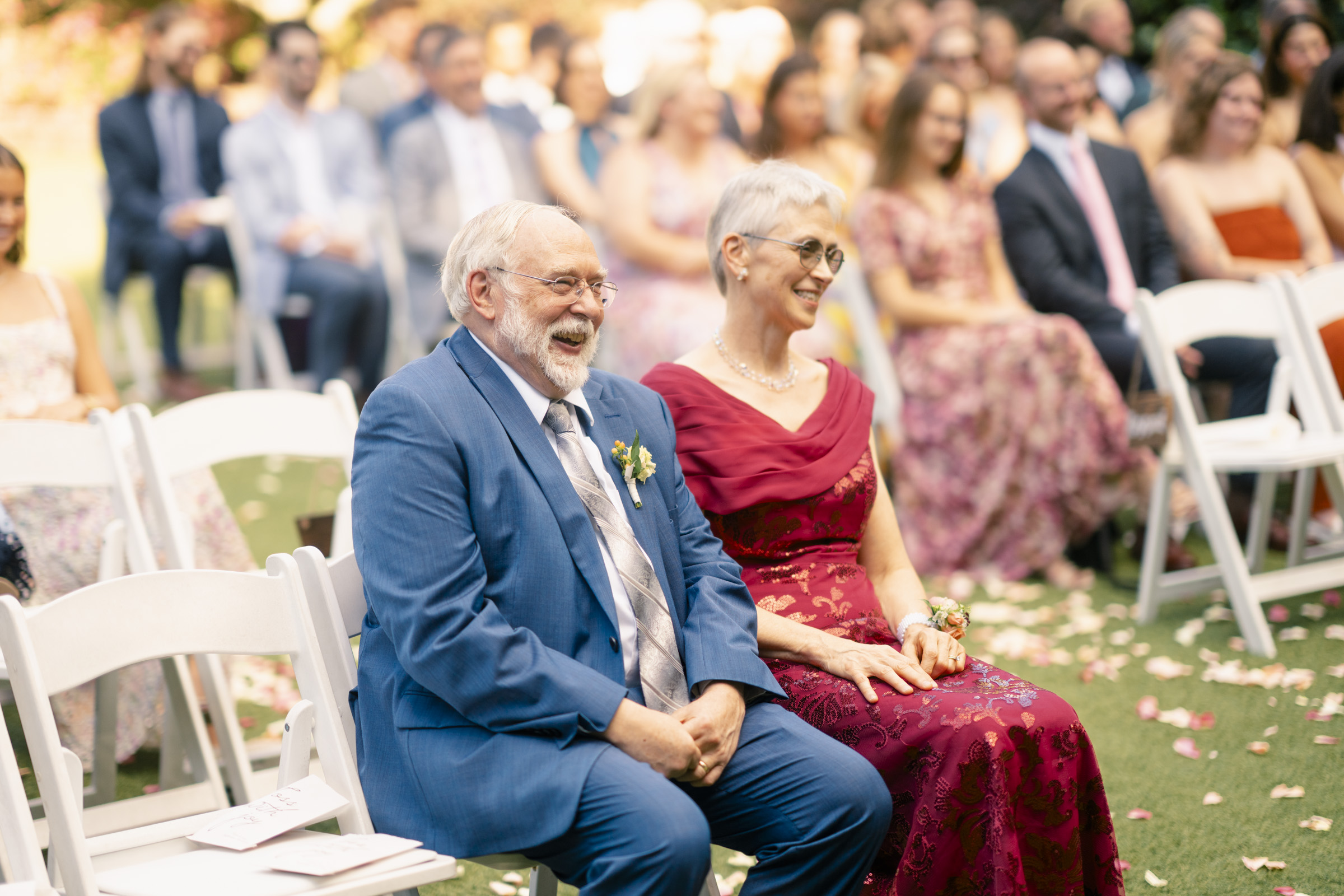 An elderly couple, dressed formally, sit on white chairs outdoors, smiling and laughing. The man wears a blue suit, and the woman is in a red dress. They are surrounded by other seated guests, blurred in the background. Blossom petals lay on the grass.