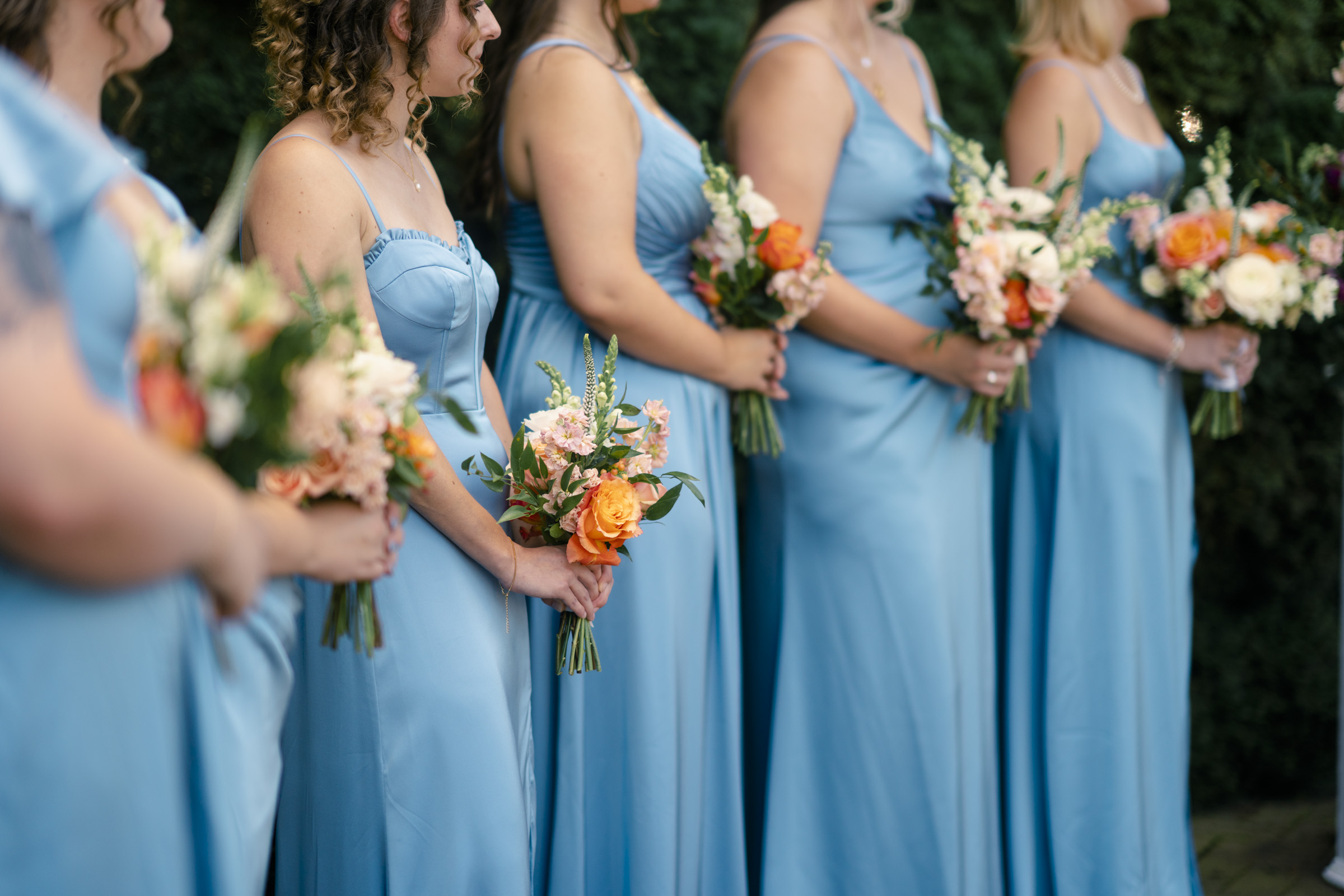 A group of people in light blue dresses stand in a line, each holding a bouquet of flowers with orange and pink hues. They are facing to the right, and greenery is visible in the background.