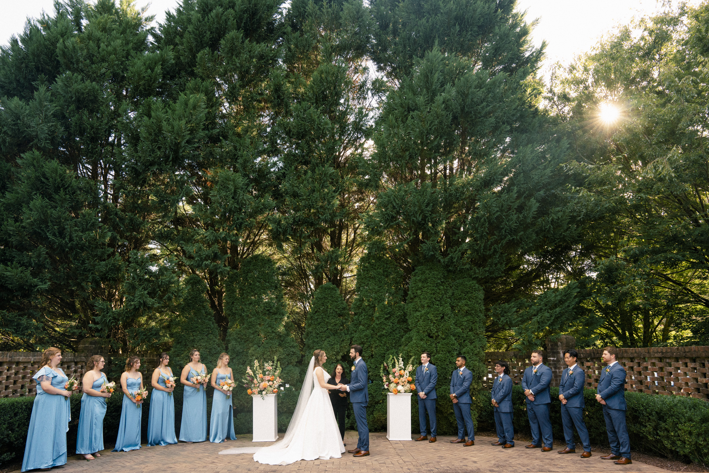 A couple stands at an outdoor wedding ceremony under tall trees. Bridesmaids in light blue dresses and groomsmen in dark suits line both sides. The sun peeks through the trees, casting a warm glow on the scene.