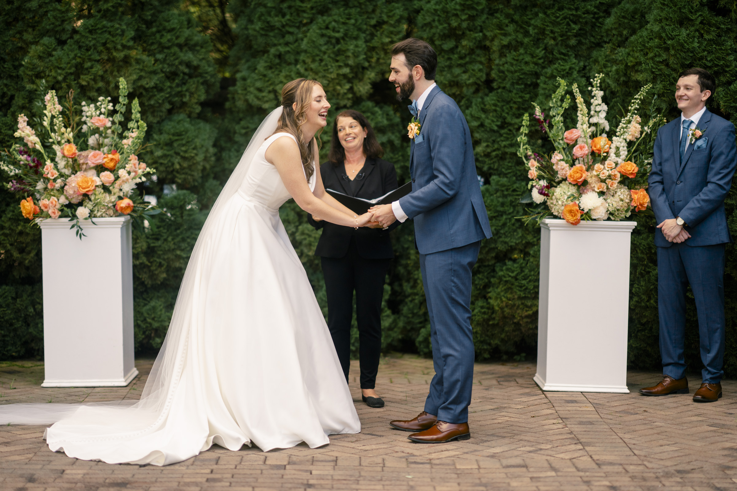 A bride and groom hold hands and smile at each other during their outdoor wedding ceremony. They stand in front of a celebrant, with floral arrangements on either side. The groom wears a blue suit, and a smiling man in a matching suit stands nearby.