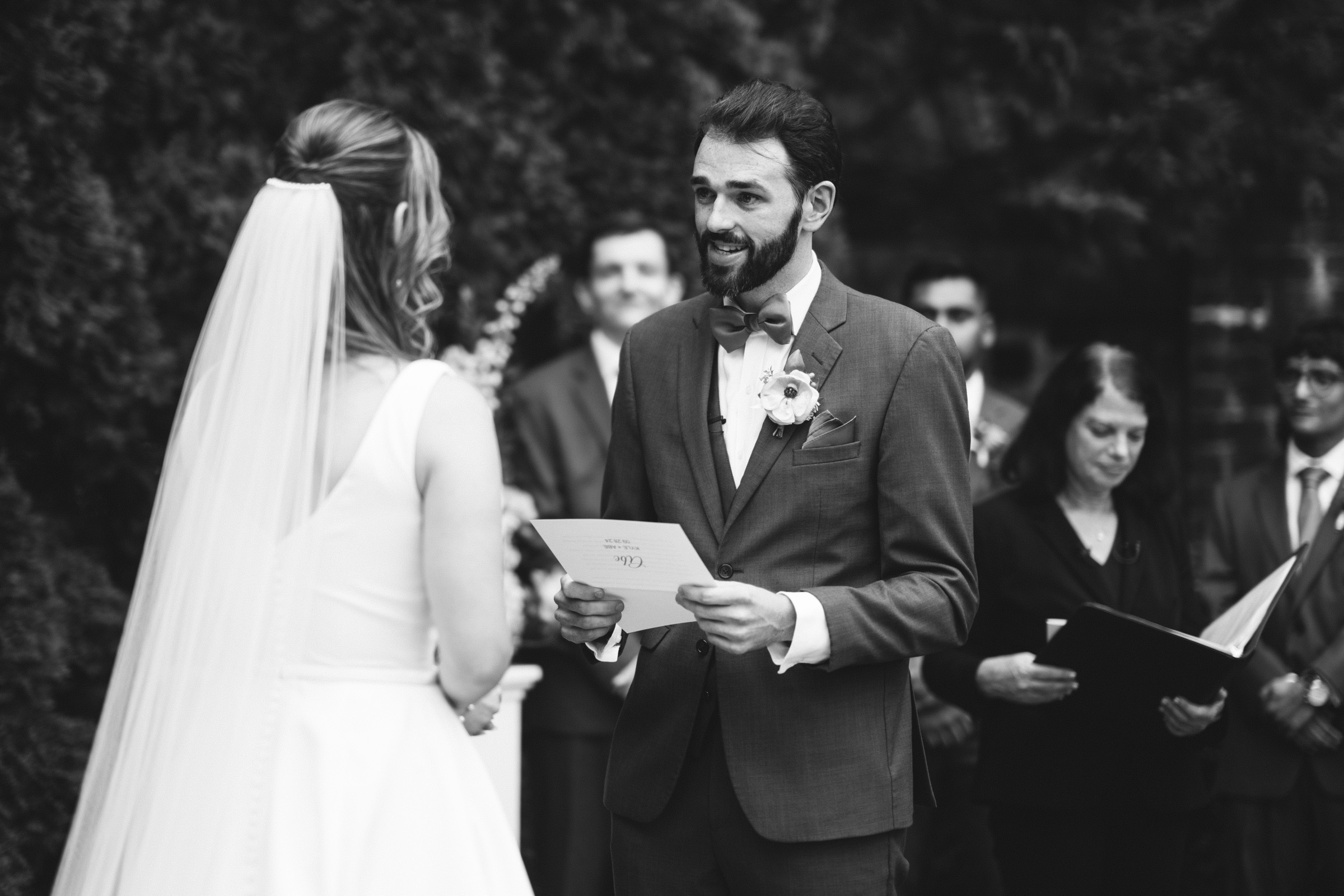 A black and white photo of a bride and groom at their wedding ceremony. The groom, holding papers, is speaking to the bride. The bride is facing him, wearing a veil and dress. A few guests and an officiant with a book are visible in the background.