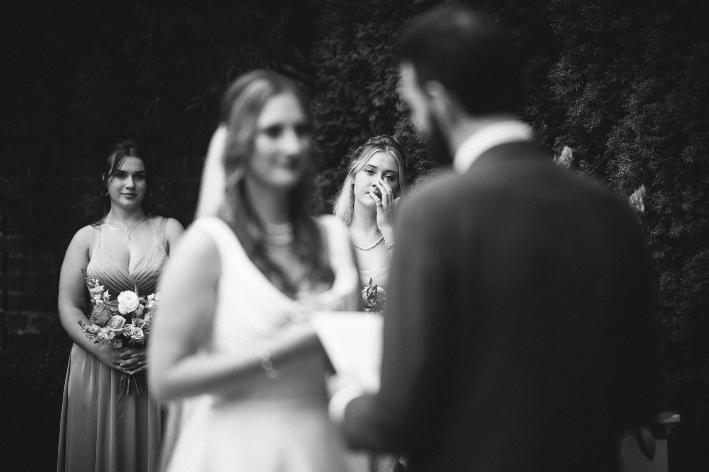 A black and white photo of a wedding ceremony. The foreground shows a bride and groom facing each other. In the background, two bridesmaids, with one touching her face, are standing near a tree. The focus is on the emotional expression of the bridesmaid.