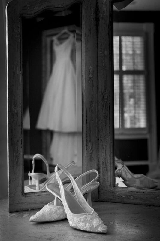 Black and white image of a pair of lace bridal shoes in front of an antique mirror. A blurred wedding dress is reflected in the mirror, hanging in the background near a window with blinds.