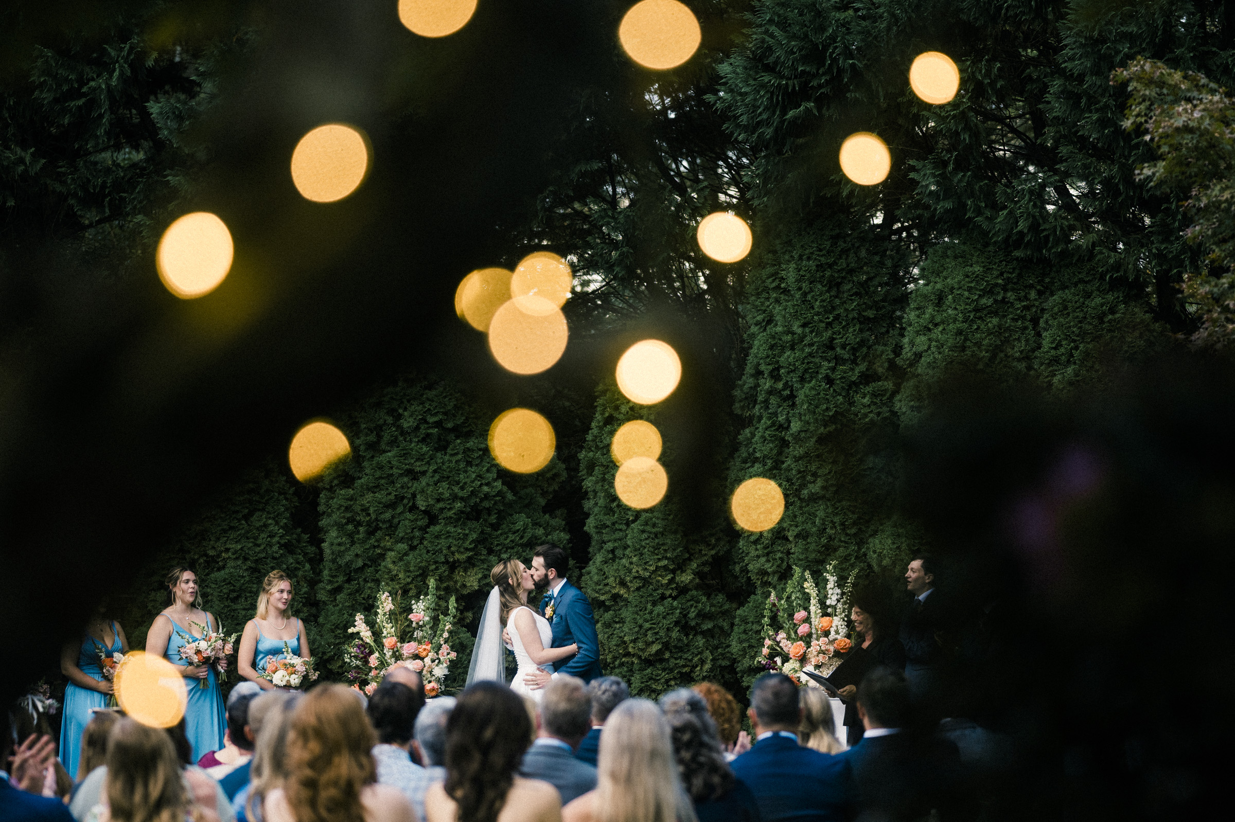 A couple kisses at an outdoor wedding ceremony. Bridesmaids in blue dresses stand nearby, with floral arrangements in the background. Blurred string lights create a bokeh effect above the seated guests. Dense greenery surrounds the scene.