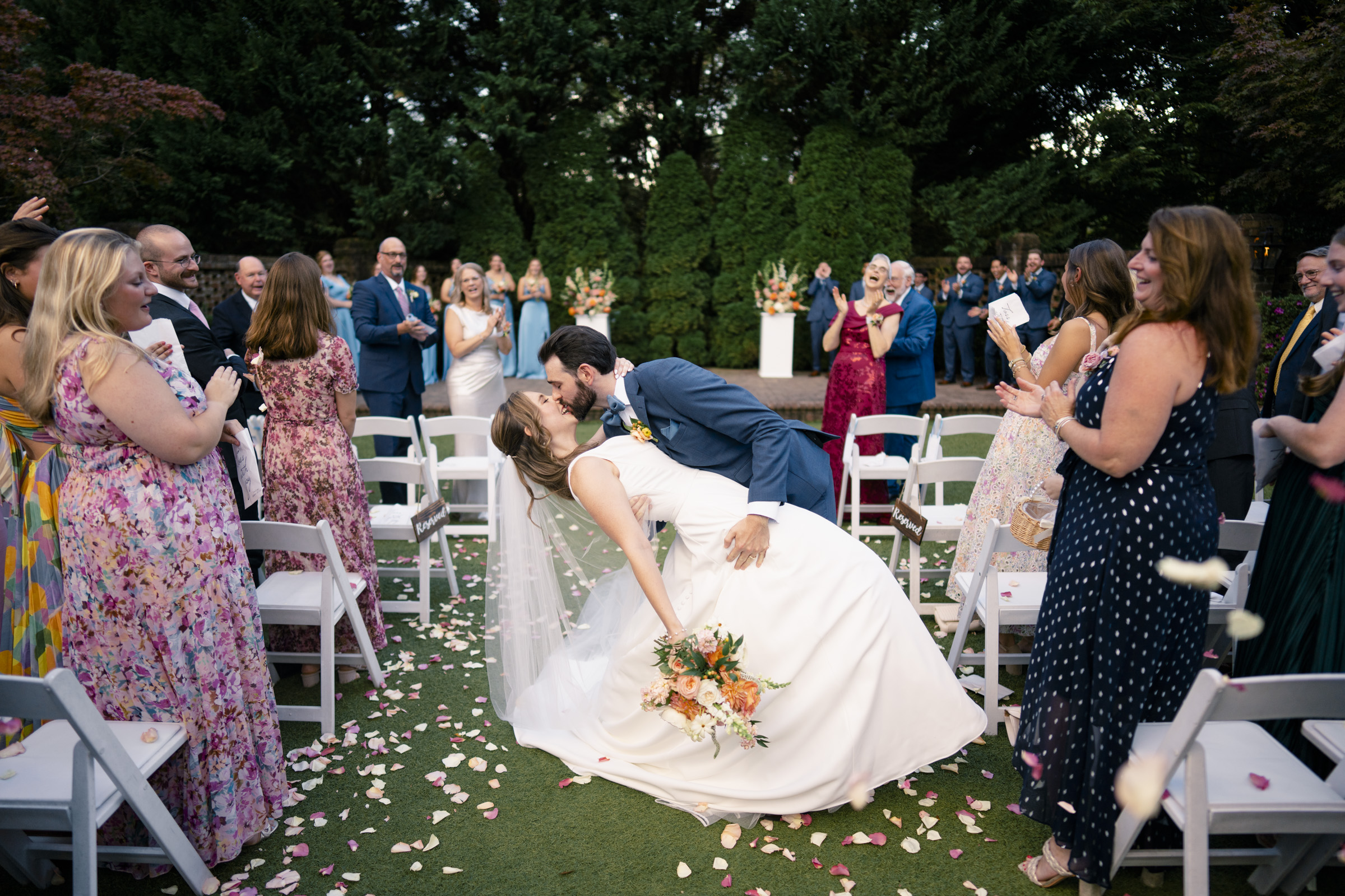 A bride and groom share a kiss in an outdoor setting while guests clap and cheer around them. The bride is holding a bouquet of flowers, and rose petals are scattered on the grass. The guests are dressed in formal attire.