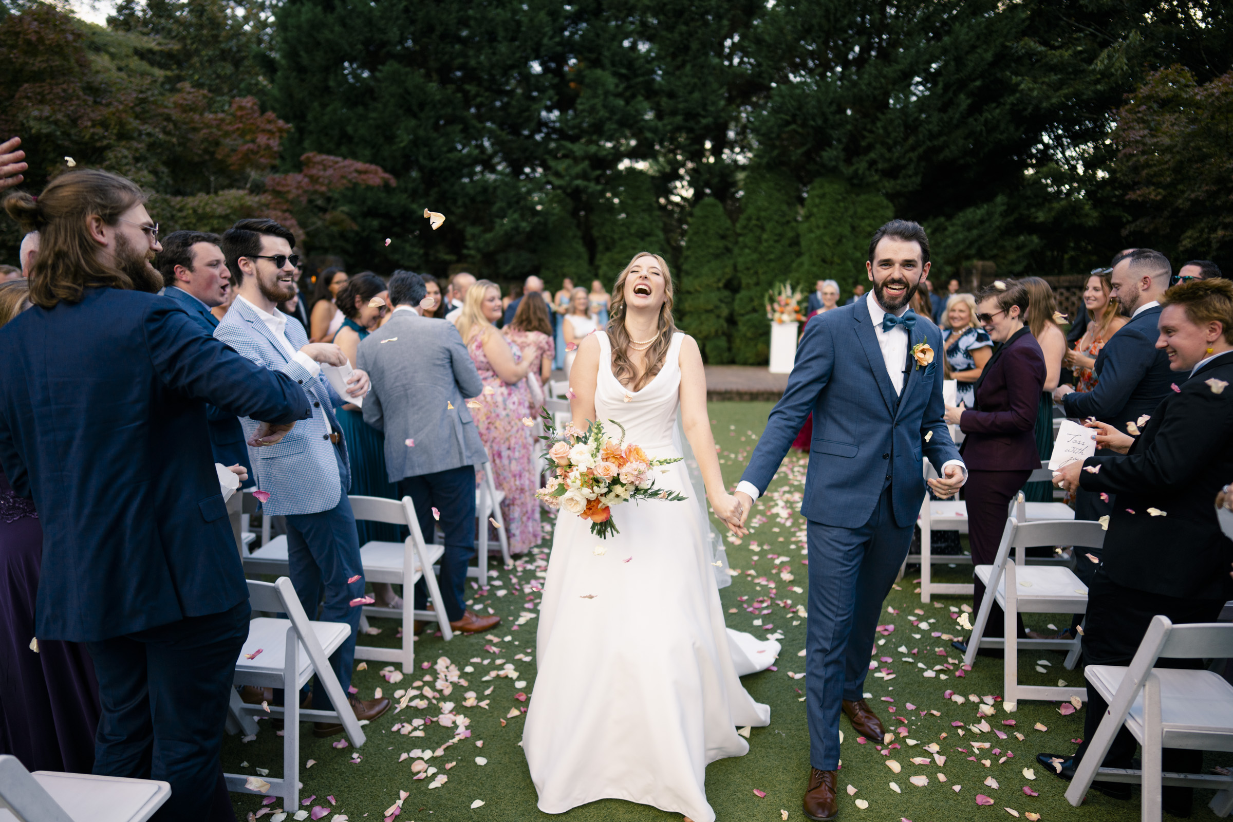 A joyful couple walks down an outdoor aisle after their wedding ceremony. The bride, in a white dress, holds a bouquet and laughs, while the groom, in a blue suit, smiles. Guests on either side clap and toss petals.