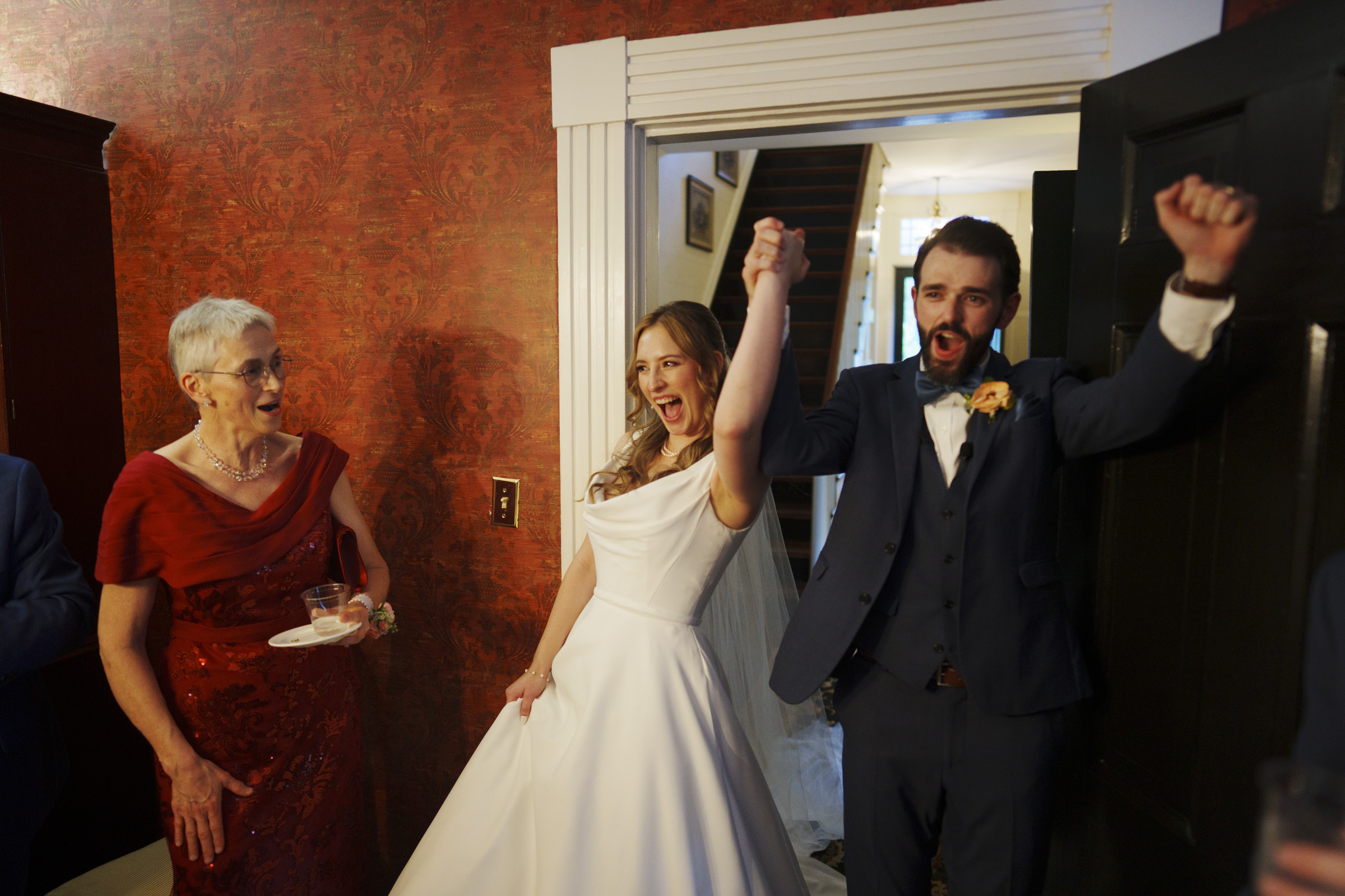 A bride and groom celebrate joyfully, holding hands and raising their arms. The bride wears a white gown, and the groom is in a blue suit. An older woman in a red dress stands nearby, smiling and holding a drink. They are indoors with warm lighting.