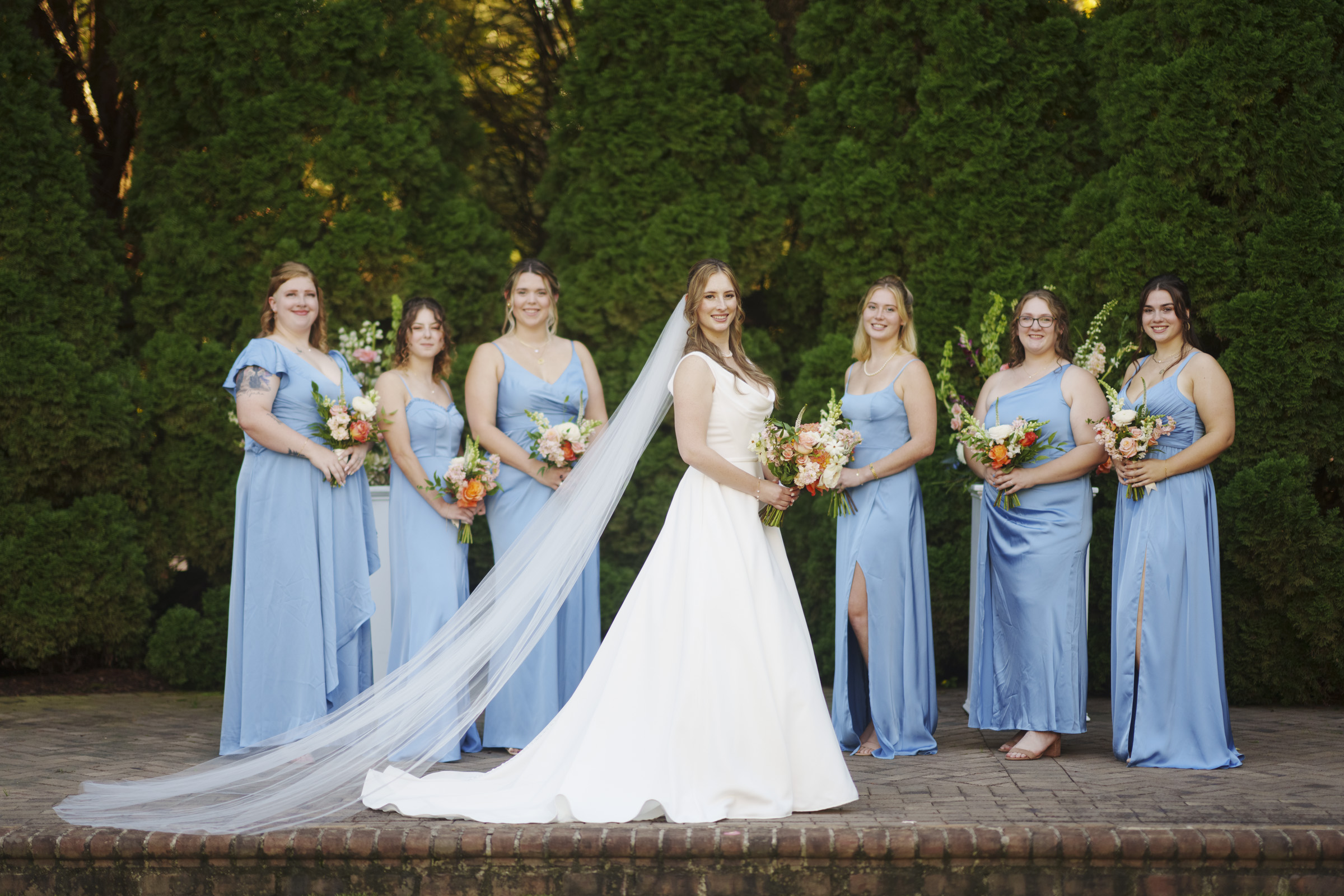 A bride in a white gown stands with six bridesmaids in light blue dresses, holding bouquets. They are outdoors with green foliage in the background. The brides long veil flows to the side.