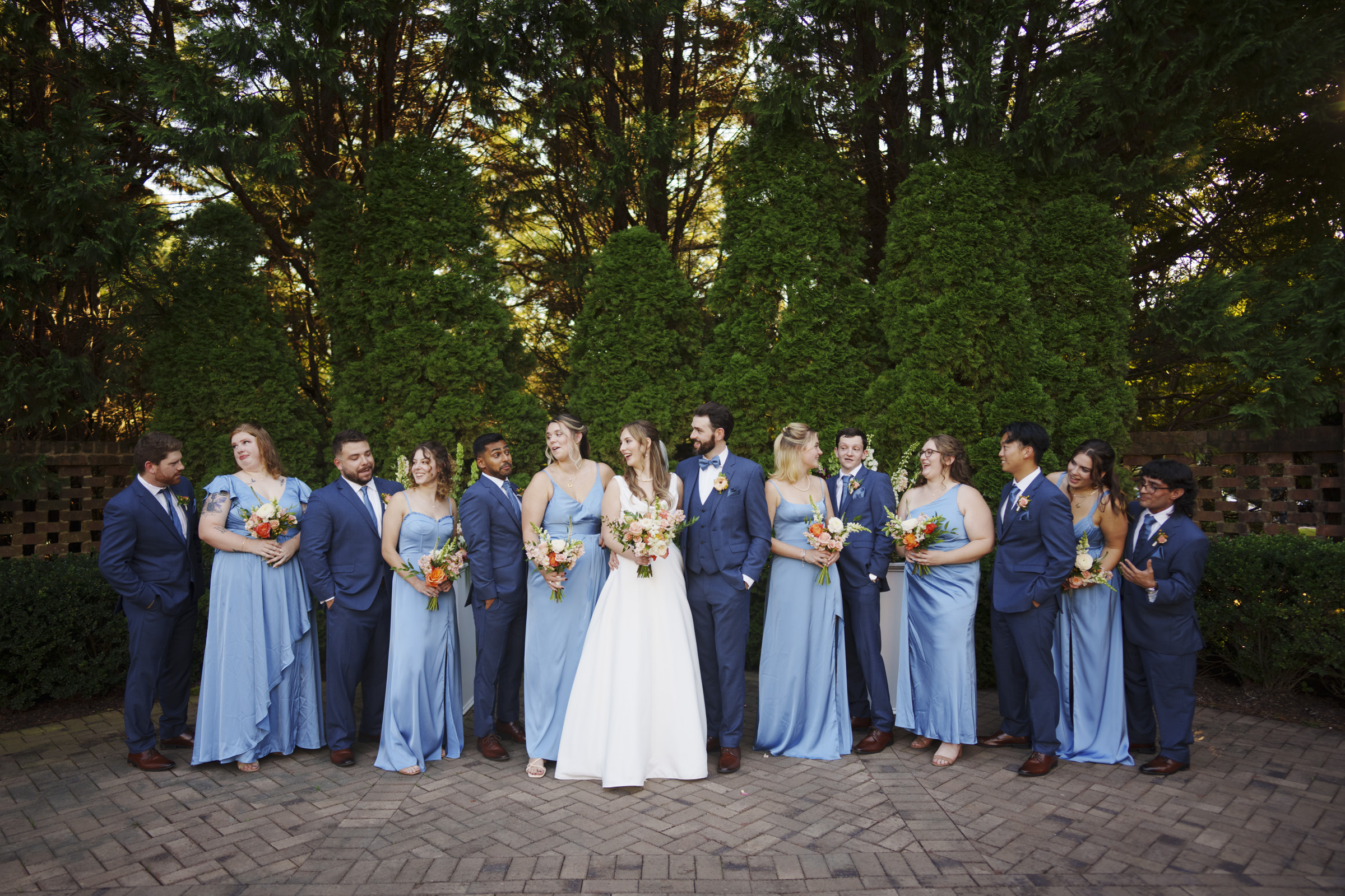 A wedding party poses outdoors in front of tall green hedges. The group includes a bride in a white dress and groom in a dark suit, surrounded by bridesmaids in light blue dresses and groomsmen in dark suits. Everyone is smiling.