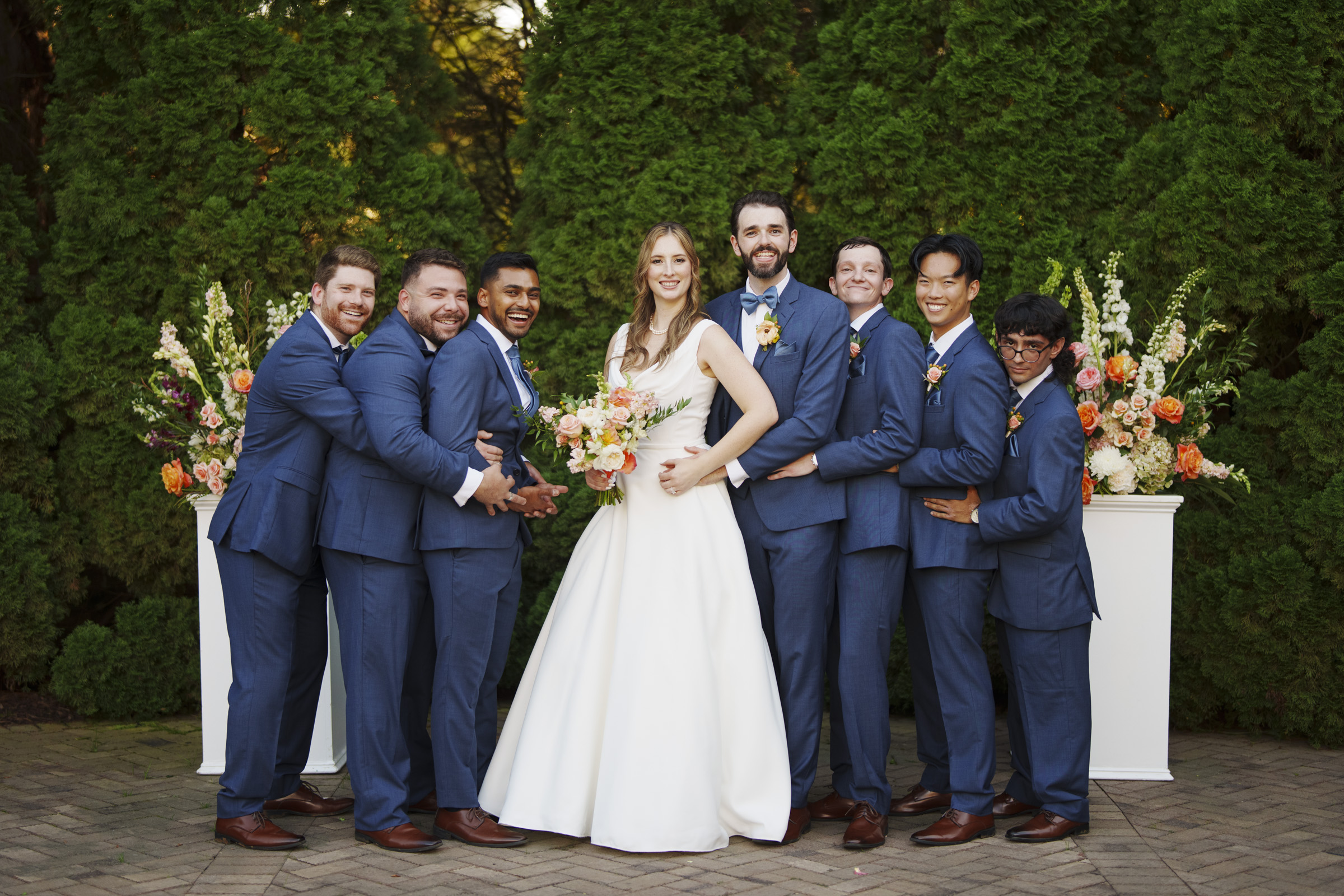 A bride in a white gown stands with six groomsmen in matching blue suits. They are all smiling and standing close together in front of lush greenery and floral arrangements.