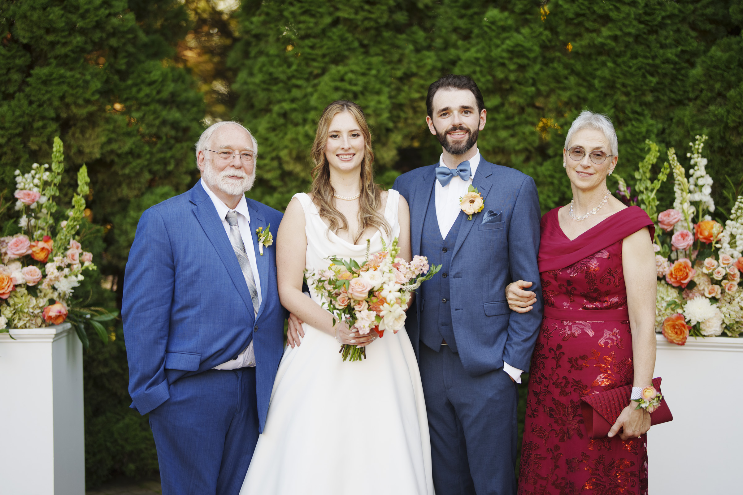 A bride and groom stand between an older man in a blue suit and an older woman in a red dress. The bride holds a bouquet of flowers. They are outdoors with lush greenery and floral arrangements in the background.