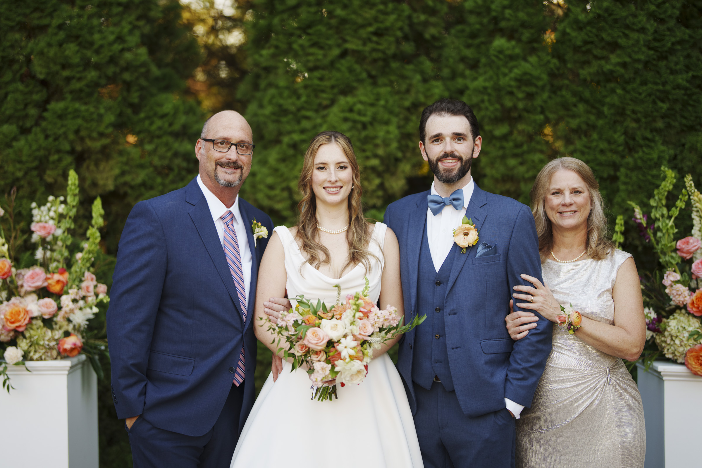 A bride and groom stand smiling with two older adults, likely parents. The group is posed in front of lush greenery with floral arrangements. The groom wears a blue suit and bow tie, the bride in a white gown holding a bouquet.