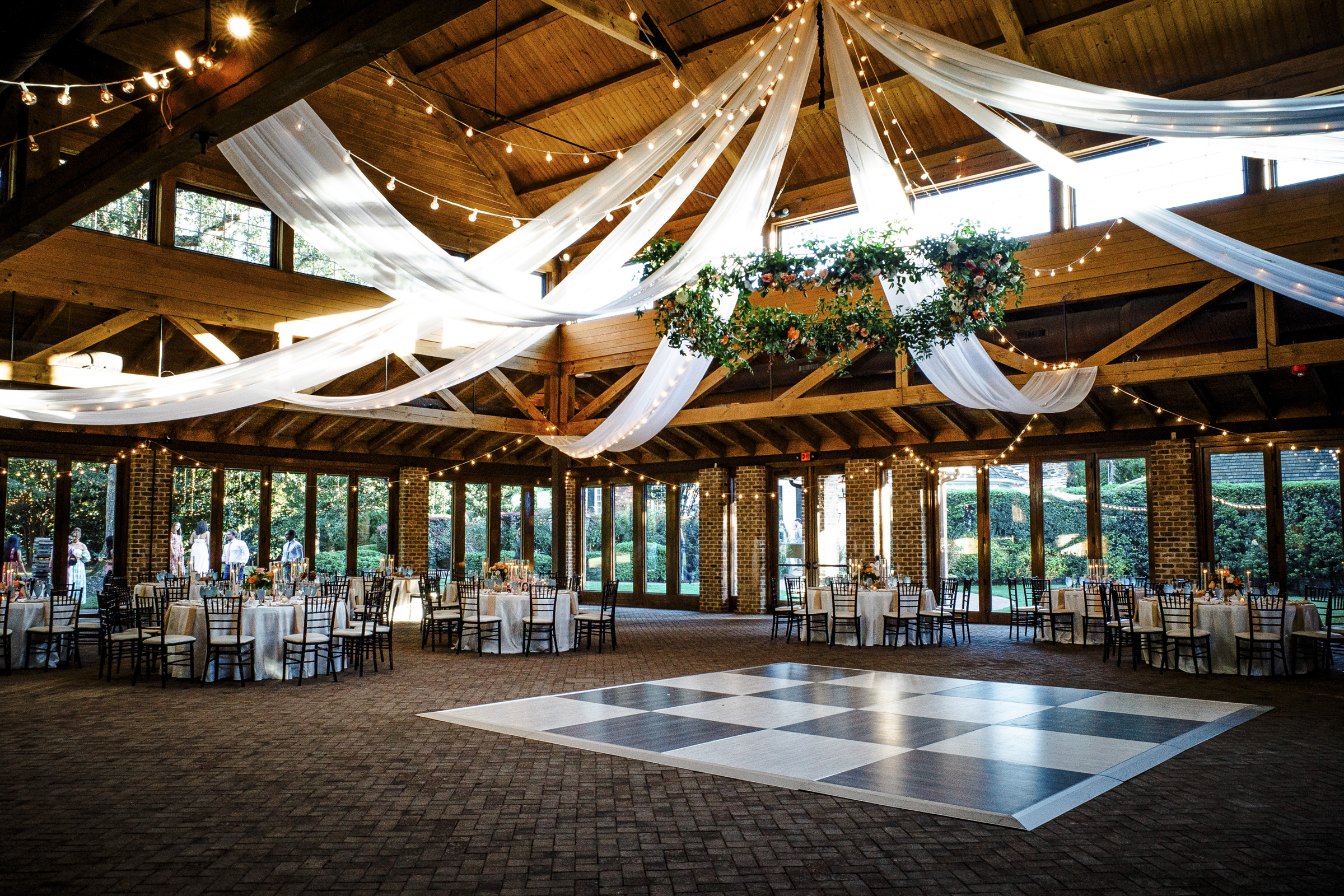 A spacious wedding venue with a wooden ceiling, draped white fabric, and string lights. Tables with white tablecloths and chairs surround a large checkered dance floor. Floral arrangements hang from the ceiling. Large windows offer a view of greenery outside.