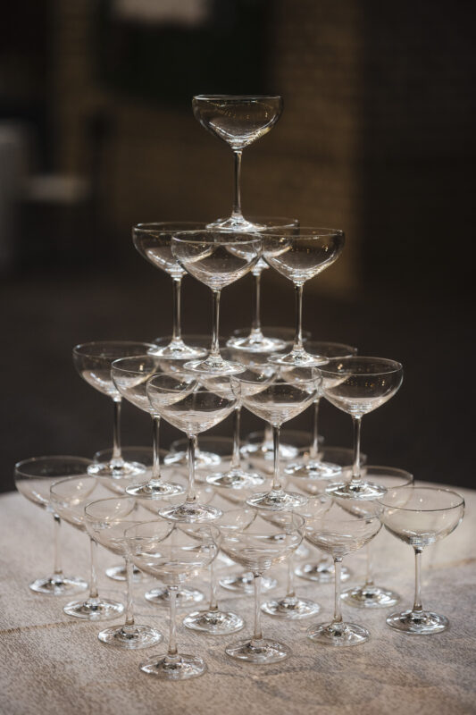 A pyramid of empty, stacked champagne glasses is displayed on a table. The glasses are arranged in a symmetrical triangular formation, creating an elegant and celebratory atmosphere against a blurred background.