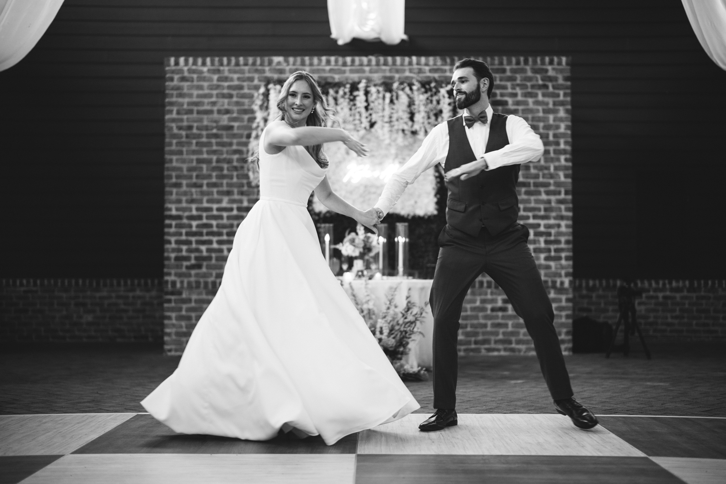 A black and white photo of a couple dancing. The woman wears a long, flowing gown, and the man is in a vest and bow tie. They are in mid-dance, smiling, with a brick wall and floral decorations in the background.