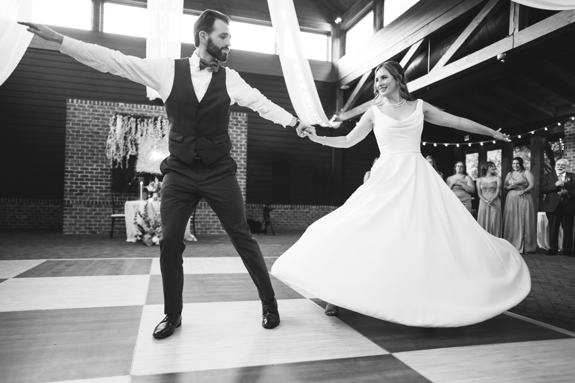 A bride and groom dance energetically on a checkered floor. The brides gown swirls as they twirl. Guests watch them in the background. The venue features wooden beams and light streams in from large windows. The scene is joyful and festive.