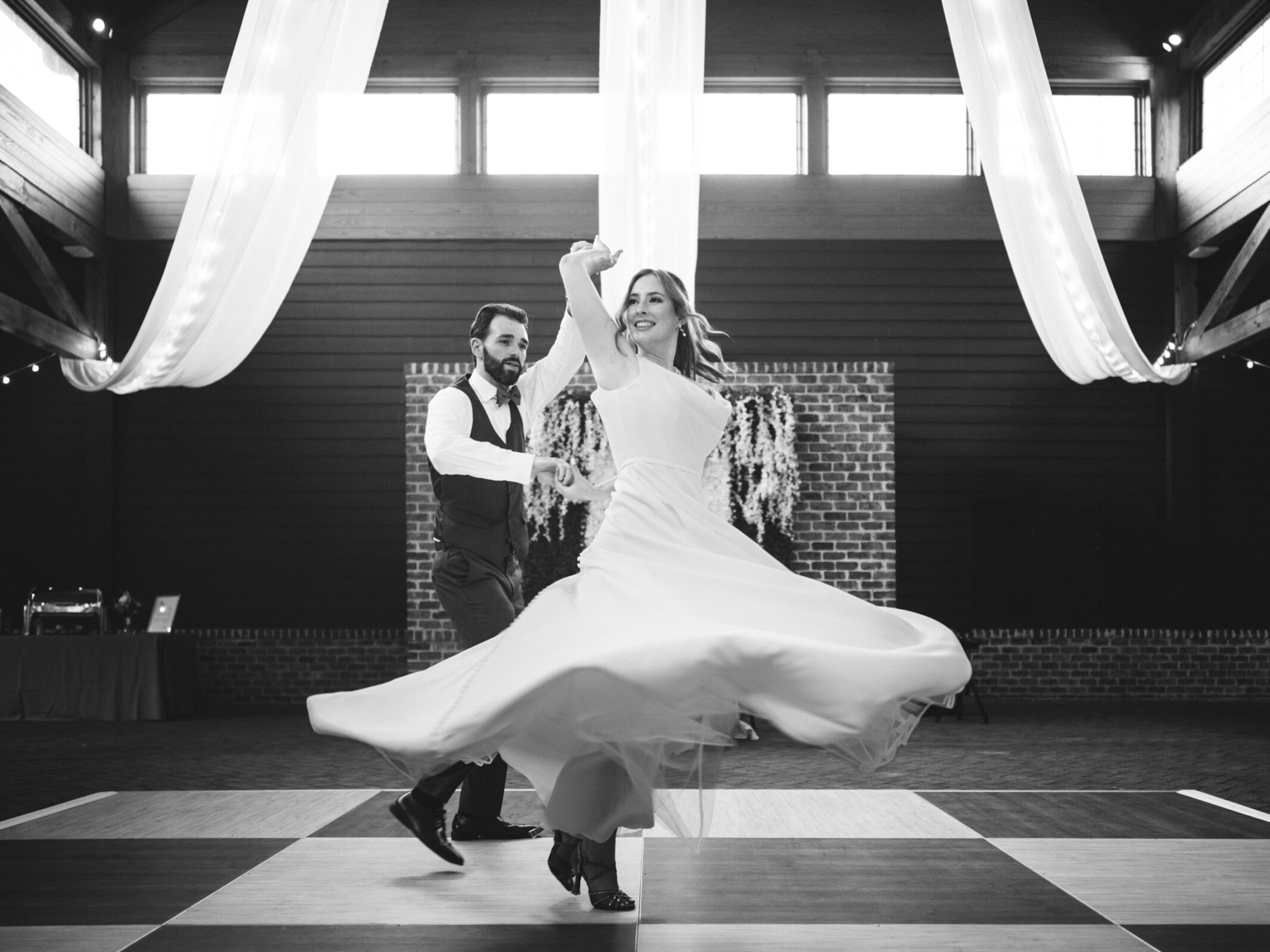 A couple dances elegantly on a checkered floor. The woman wears a flowing dress, and they appear to be twirling. Draped fabric decorates the ceiling, and a brick wall with floral arrangements is in the background. The scene is in black and white.