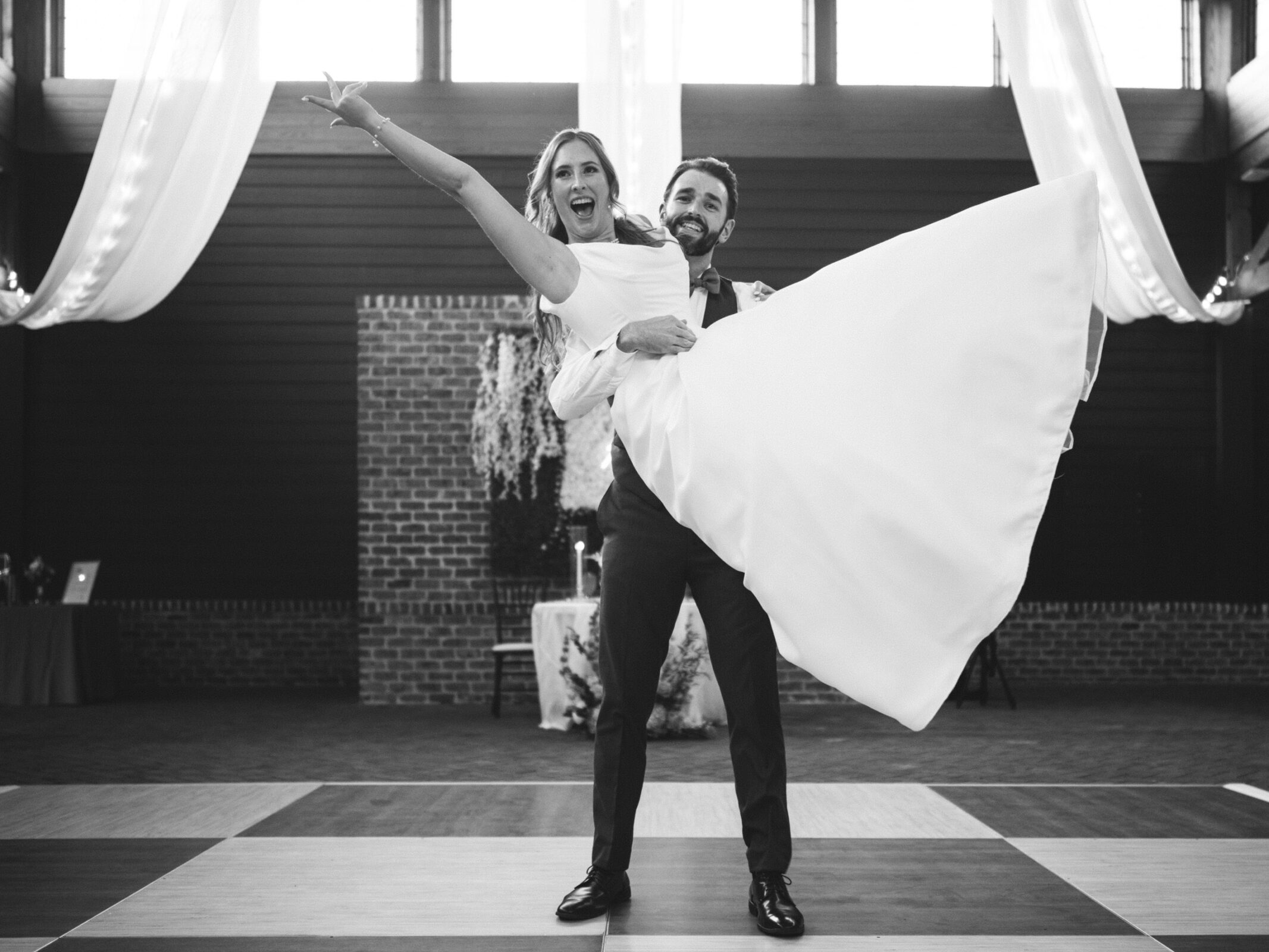 A groom playfully lifts the bride, both smiling joyfully. They are indoors, with draped curtains and warm lighting. The bride wears a long white dress, and the groom is in a suit, standing on a tiled floor.