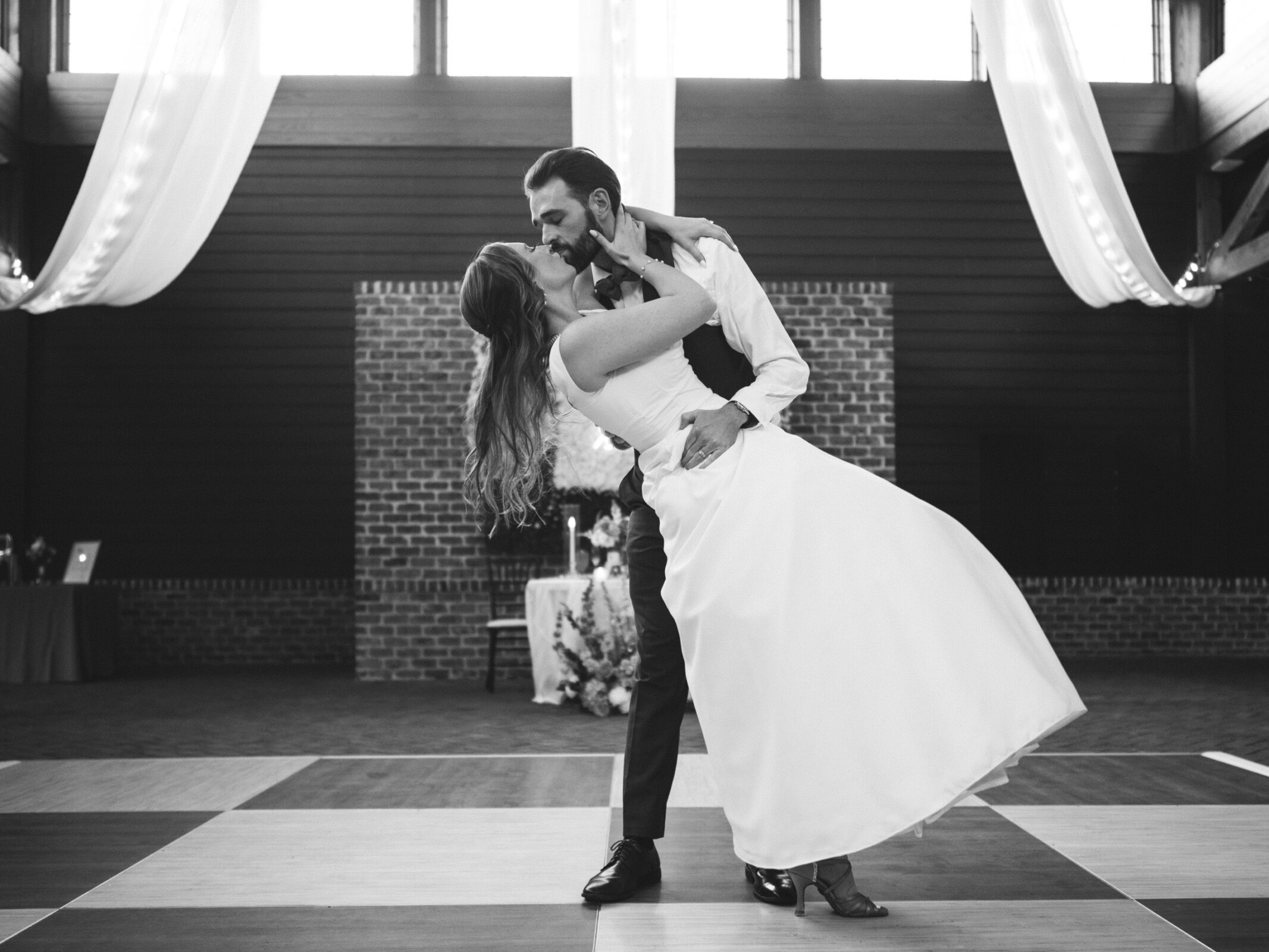 A couple is elegantly dancing in a spacious hall with draped fabrics and large windows. The groom, in a suit, dips the bride, who is in a strapless gown. They share an intimate moment on a checkered dance floor. Black and white photograph.