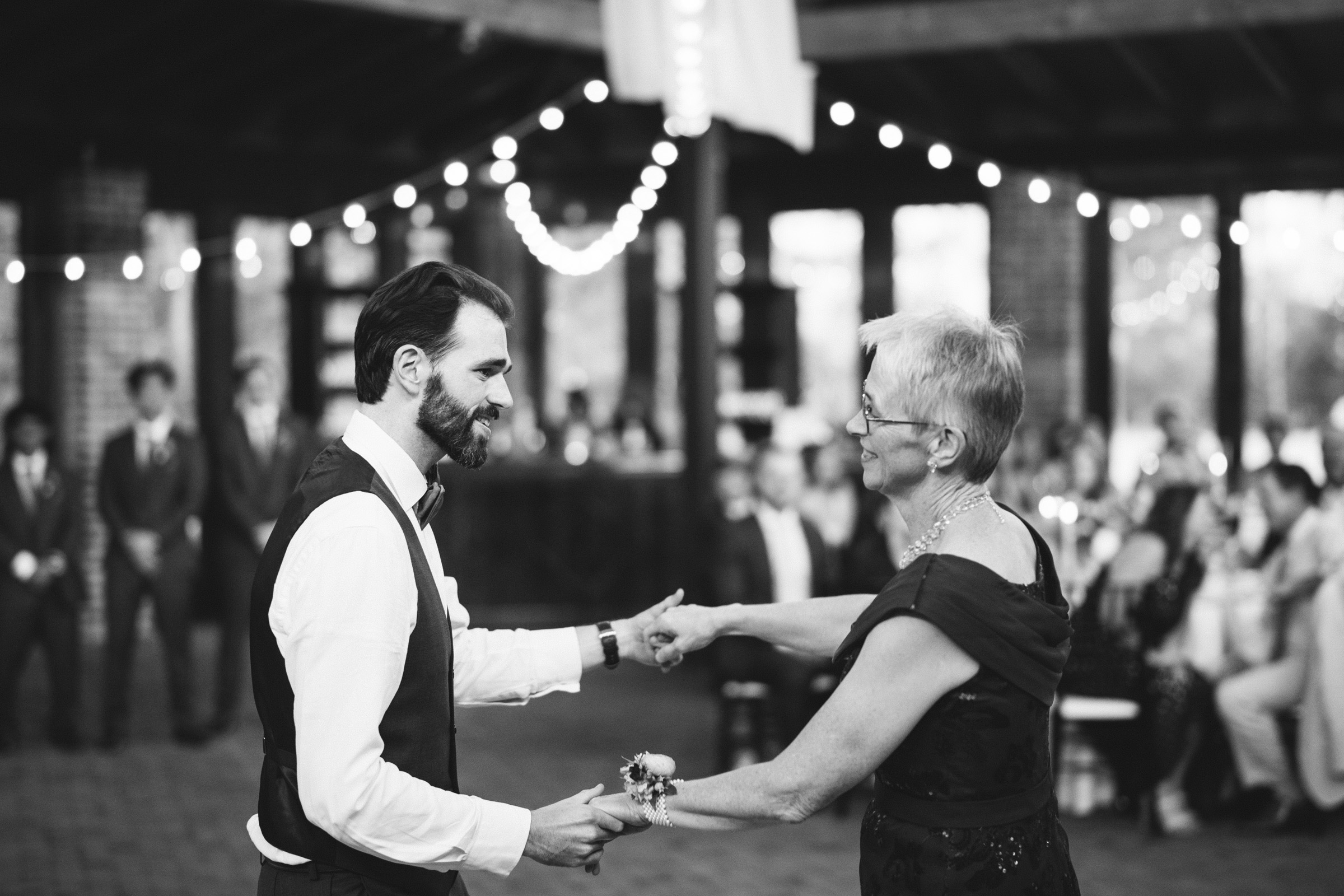 A black and white photo of a groom and an older woman, likely in a mother-son dance. They hold hands and smile at each other. Blurred background shows string lights, people seated, and a few standing.