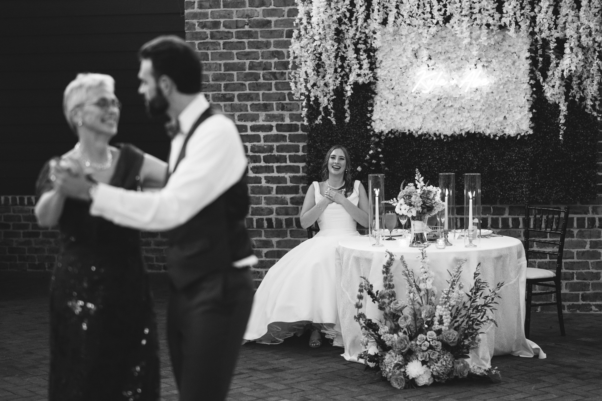 A bride sits at a decorated table, smiling and clapping. In the foreground, a man in a vest dances with an older woman in a dark dress. They are in a brick-walled venue with a lush floral backdrop. Black and white photo.