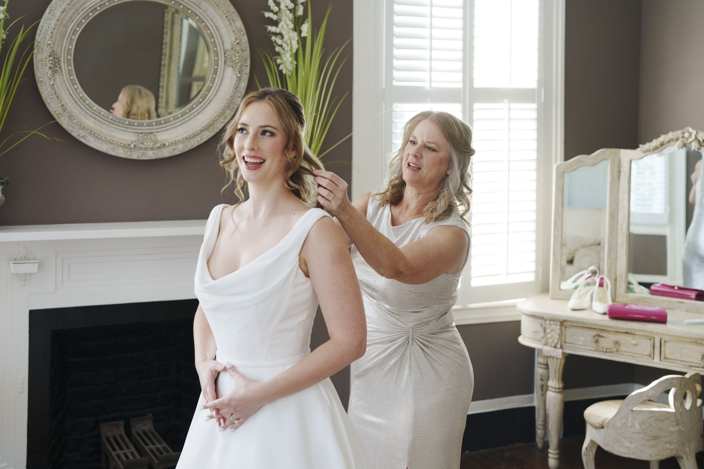 A bride in a white dress stands smiling as another woman adjusts her hair in a well-lit room with elegant decor. A vintage mirror and a vanity table are in the background, along with a window with shutters.