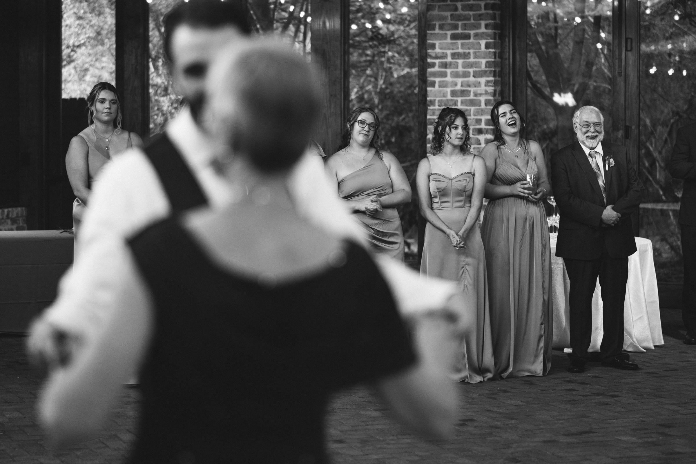 A black and white photo of a couple dancing in the foreground. In the background, several people, including three women in dresses and a man in a suit, watch and smile. They stand against a brick wall with large windows.