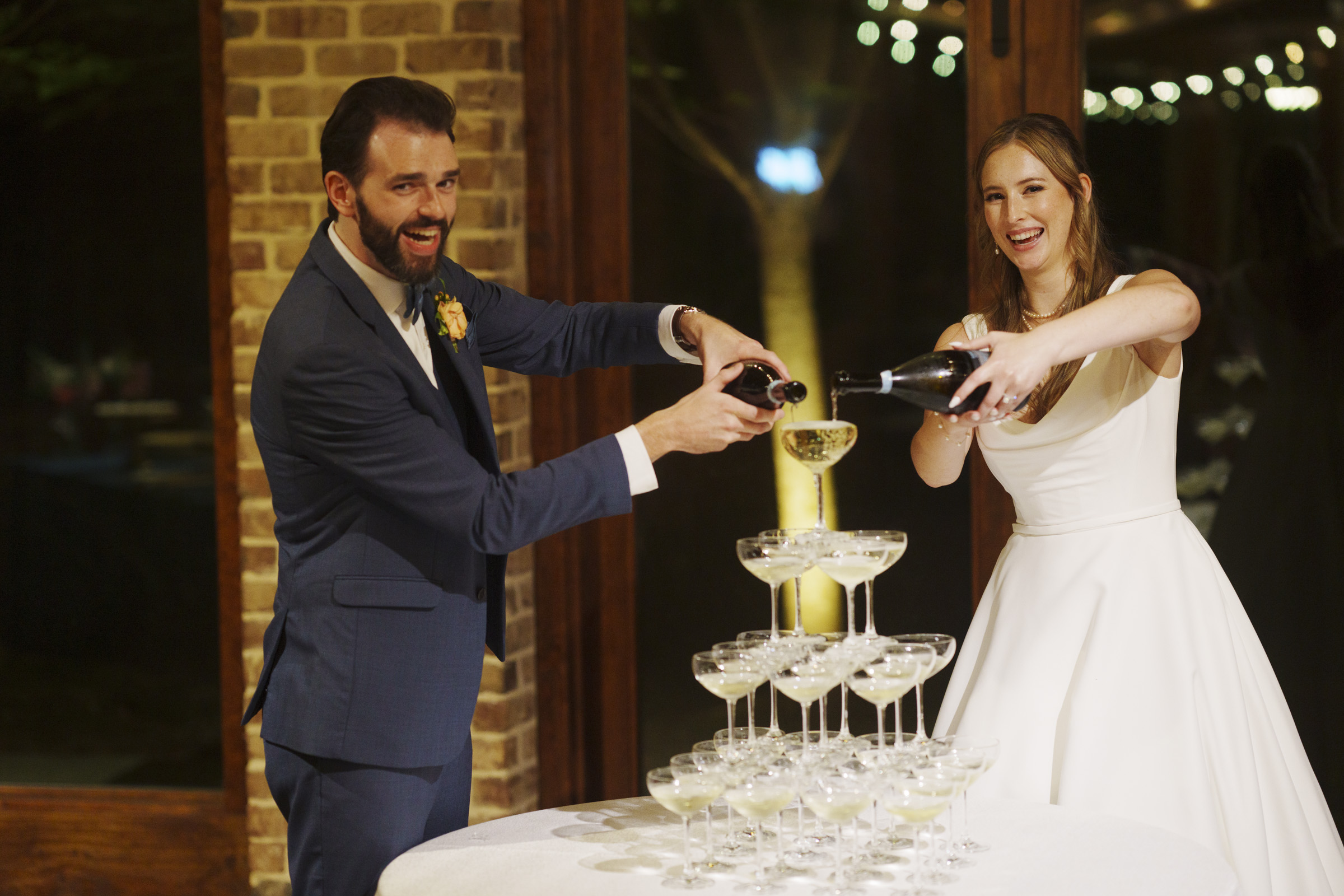 A bride and groom happily pour champagne into a towering pyramid of glasses at a wedding reception. They are dressed formally; the bride in a white gown and the groom in a blue suit. String lights and a brick wall are visible in the background.