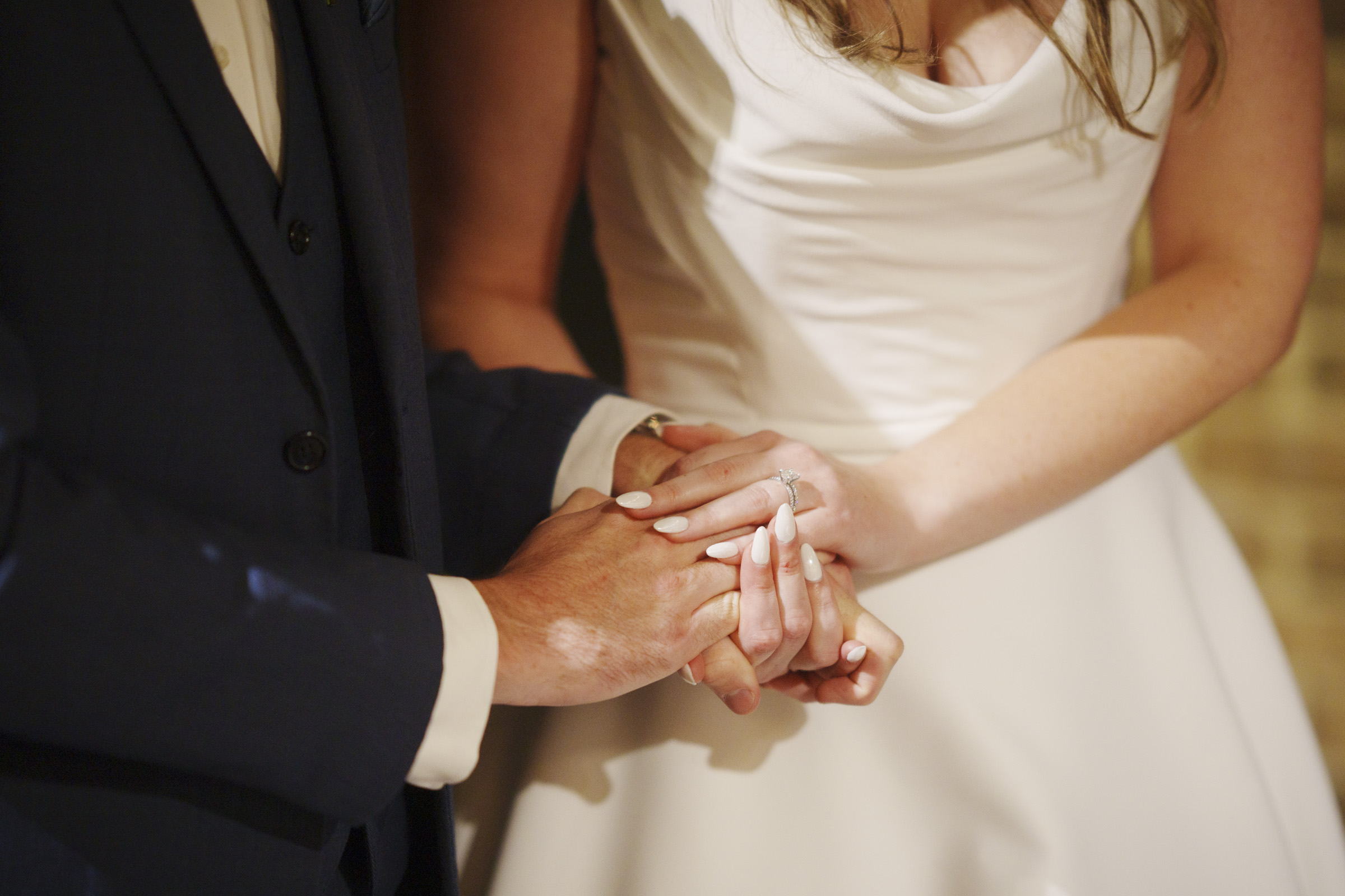 A couple holds hands during a wedding ceremony. The bride is wearing a white gown, and her engagement ring is visible. The groom is in a dark suit. Their hands are interlocked, showing a moment of connection and commitment.